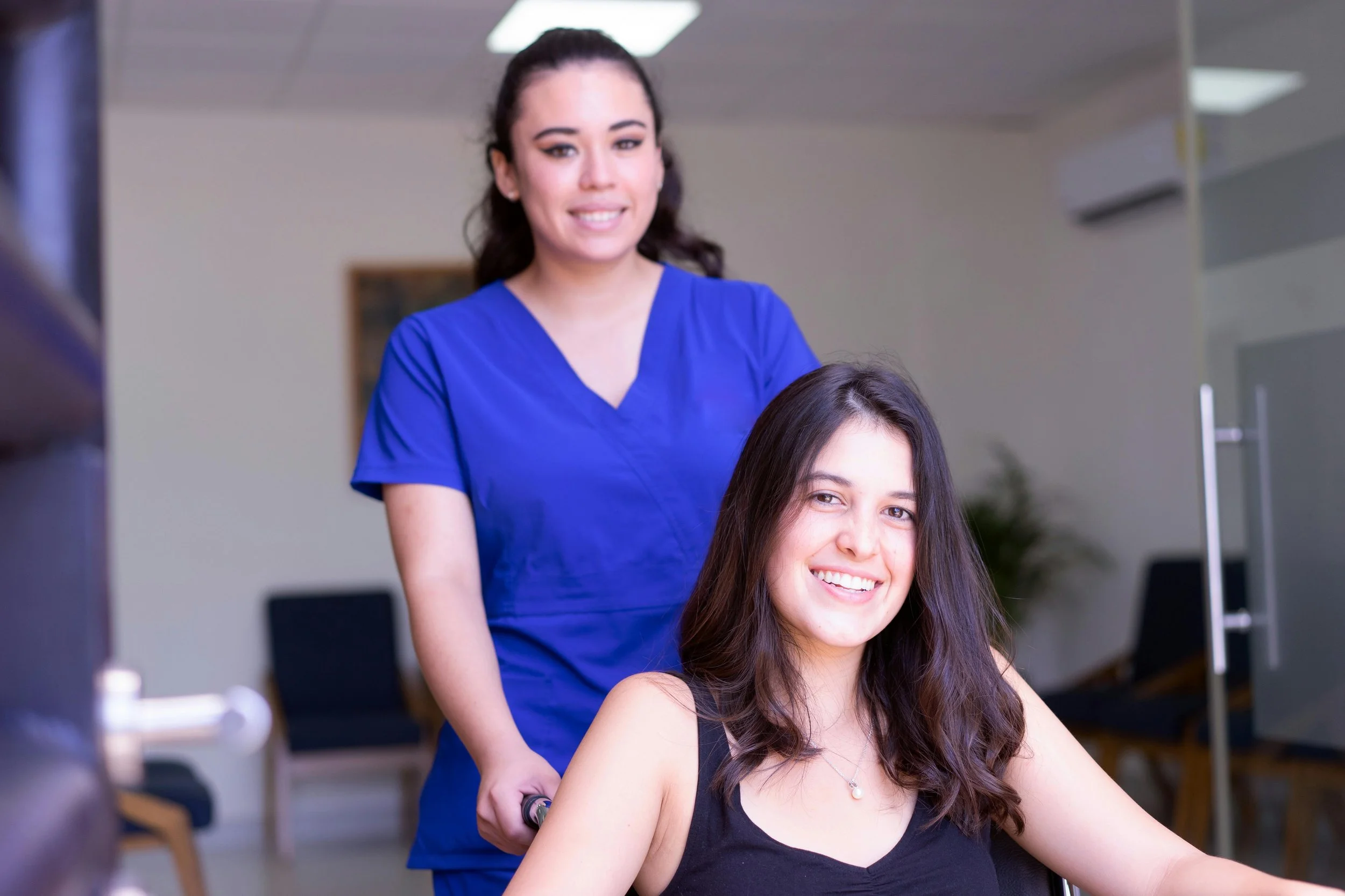 A woman with long dark hair and a black tank top smiling as she gets a massage from a female therapist wearing blue scrubs in a wellness center.