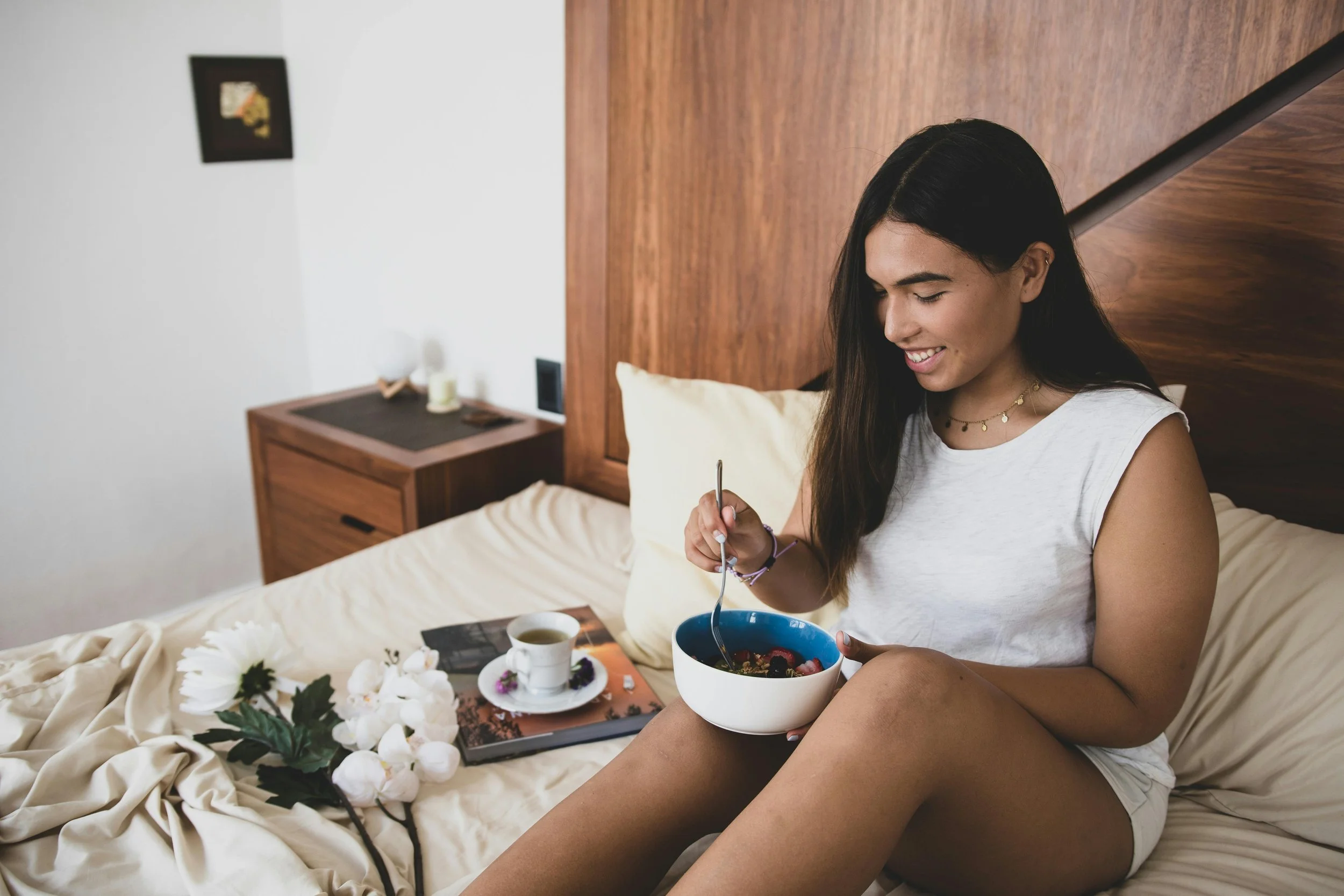 A woman sitting on a bed, smiling, holding a bowl of berries. There are flowers, a cup of tea, and a plate with food on the bed.