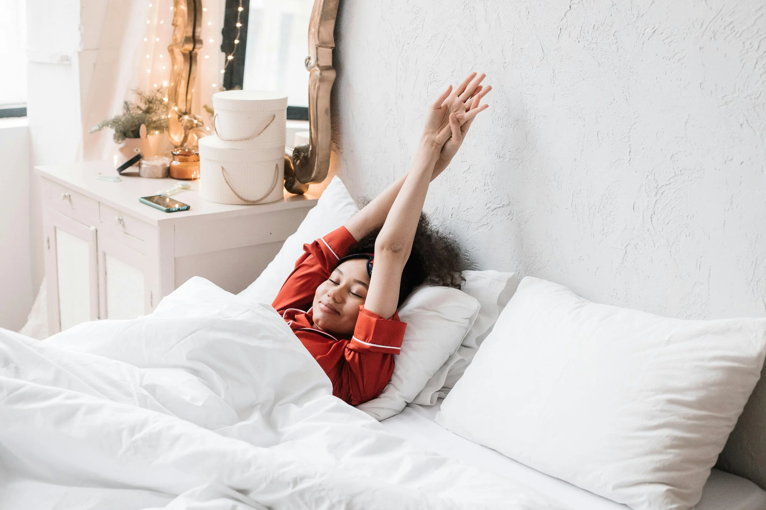 A young girl with curly hair smiling and resting in bed with her eyes closed, wearing red pajamas. She is stretching her arms above her head while lying under white bedding in a bedroom decorated with a mirror, fairy lights, a small plant, and various items on a white bedside table.