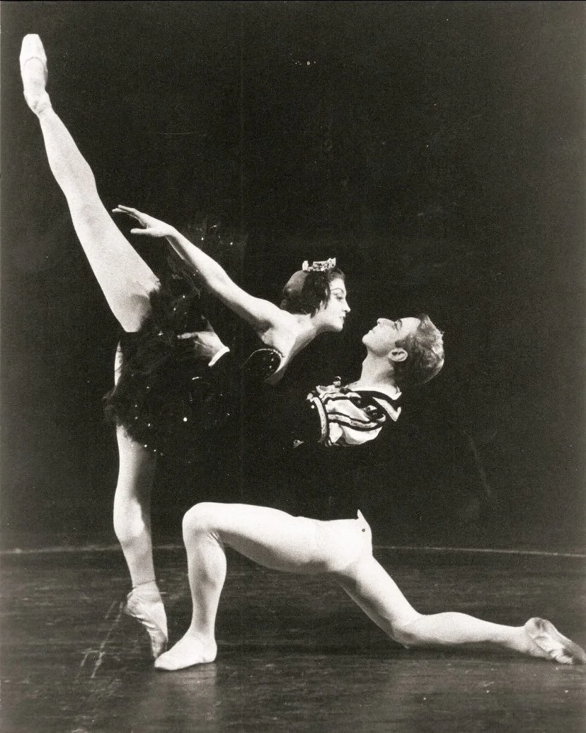 A male ballet dancer in a kneeling pose with one knee on the ground and the other leg extended backward, and a female ballet dancer in a leap with one leg extended straight up and the other bent behind her, both performing ballet on stage.