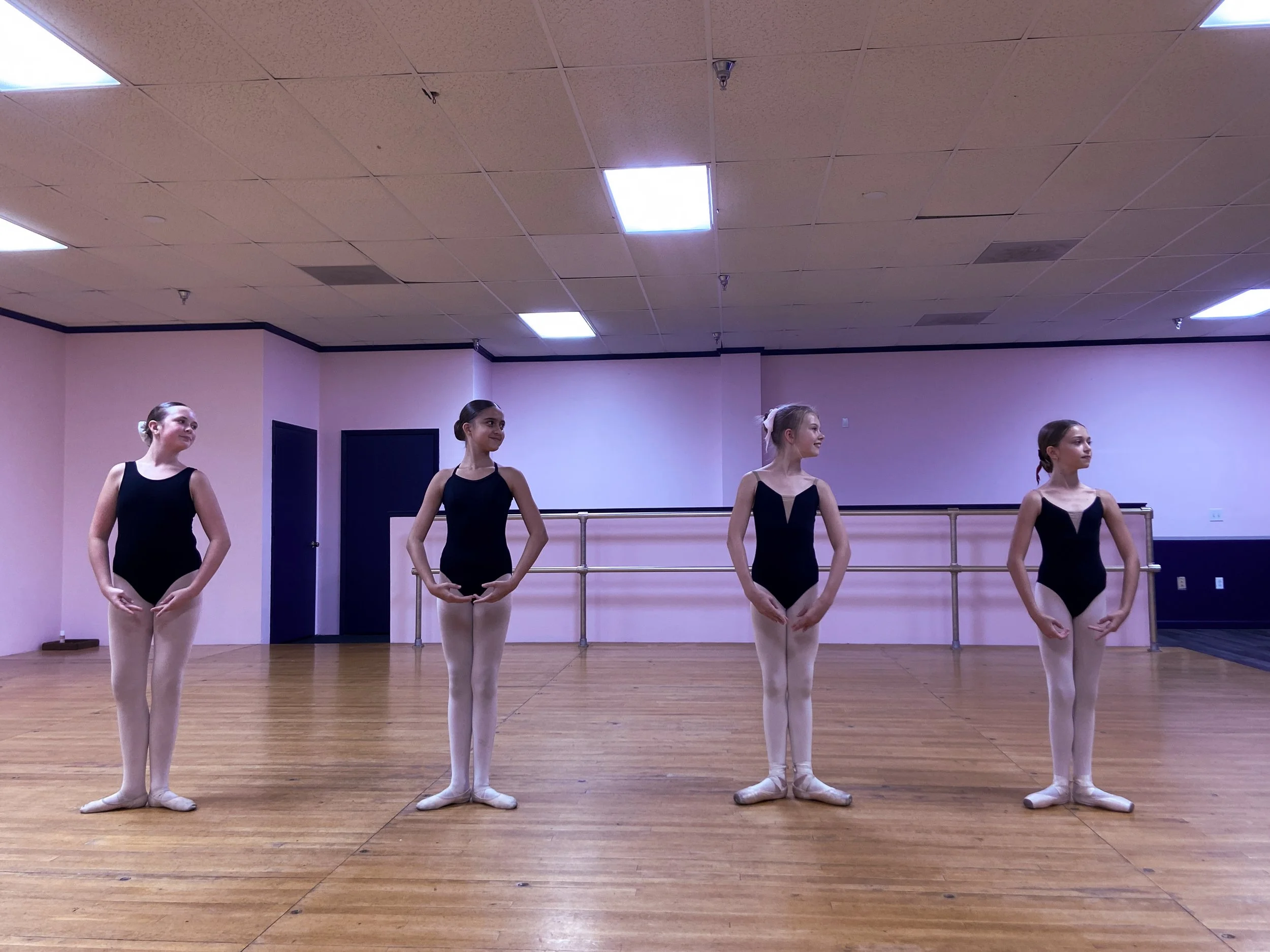 Four ballet dancers standing in a dance studio, wearing black leotards and pink tights, with their hands on their hips, looking to the side. The studio has a wooden floor, pink walls, and a ballet barre.