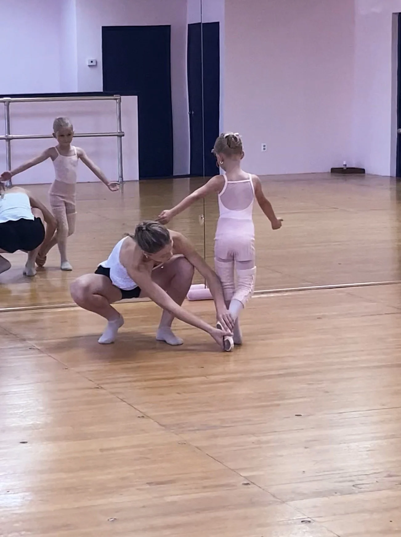 Children practicing ballet in a dance studio, with one girl in pink leotard and tights, and an instructor assisting her, while other children in ballet attire observe in the background.