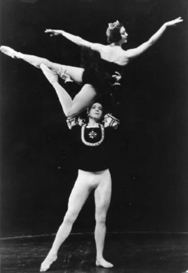 A male figure standing with one arm raised, supporting a female figure balancing on his shoulder with her arms extended outward and one leg lifted behind her. The photo is in black and white.