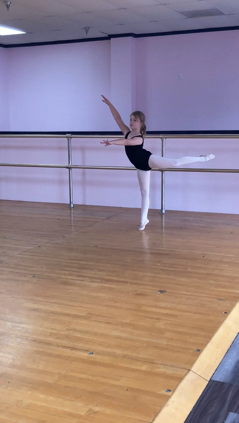 A young girl practicing ballet in a dance studio, standing on one leg and extending her other leg behind her, wearing ballet attire.