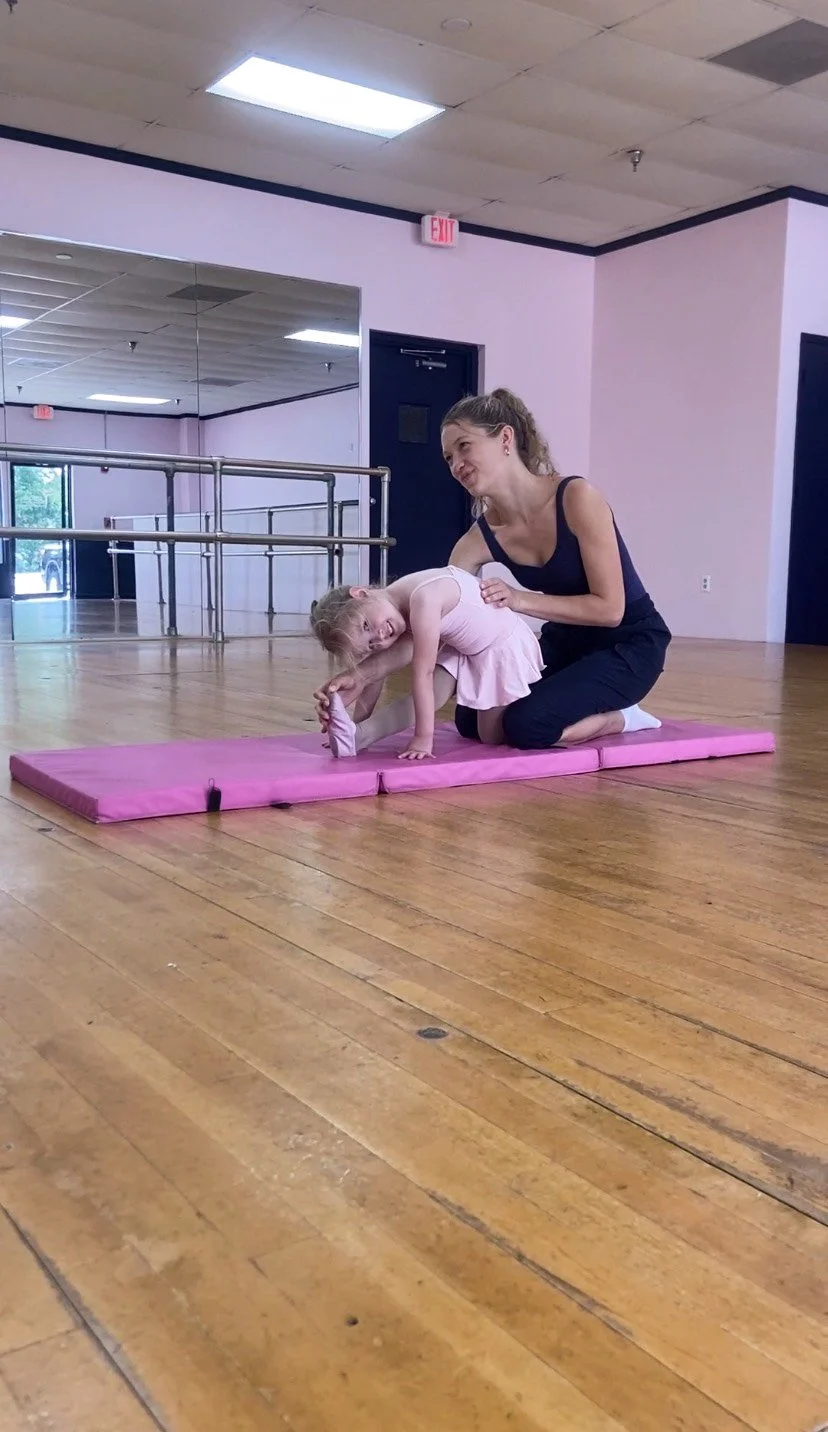 A young girl in a pink dress stretching on a pink gymnastics mat, assisted by a woman in a black tank top and pants, inside a dance studio with mirrored walls and hardwood floors.