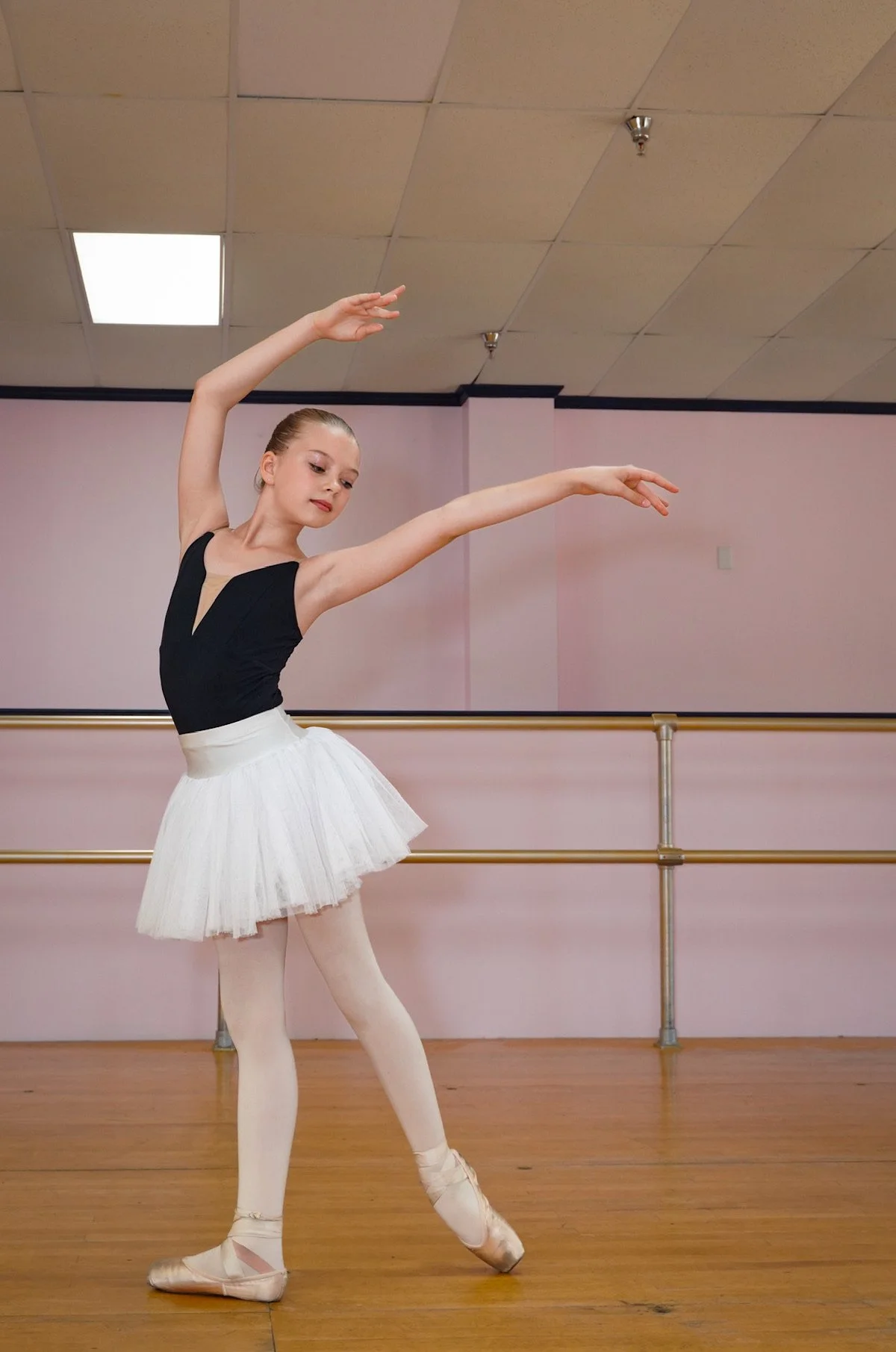 A young girl in a black ballet leotard, skirt and pointe shoes practicing ballet in a mirrored dance studio.