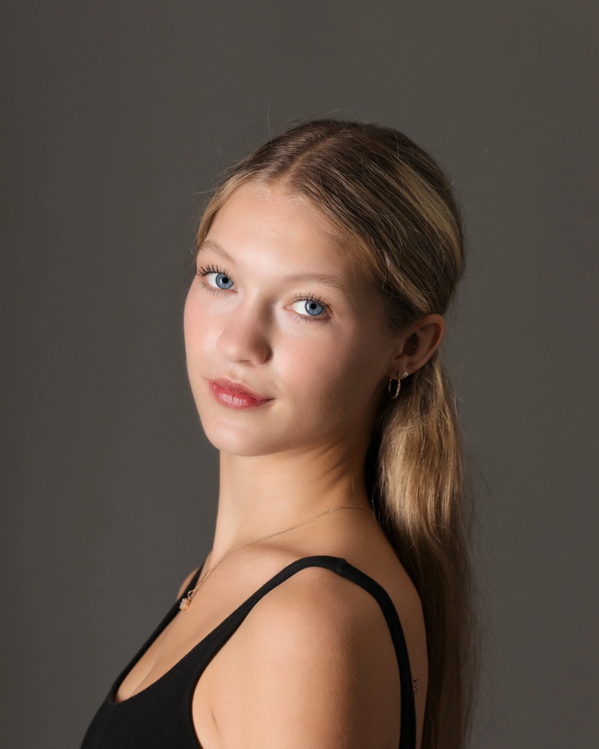 Portrait of a young woman with blue eyes, long light brown hair, and light skin, wearing a black top and gold jewelry against a gray background.