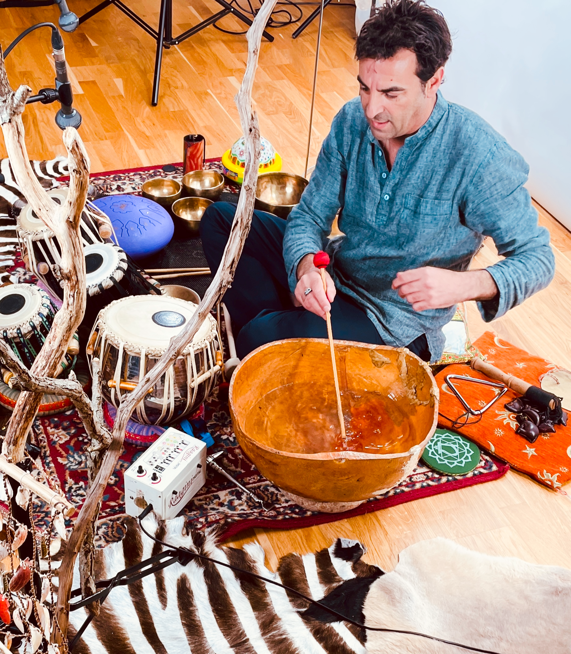 Un homme assis sur un tapis, entouré d'instruments de musique comme des bols tibétains, un tambour et une boîte à sons, en train de jouer avec un bâton. Il y a un chien blanc avec des taches noires et brunes allongé sur le tapis. La scène semble être une séance de relaxation ou de méditation musicale.