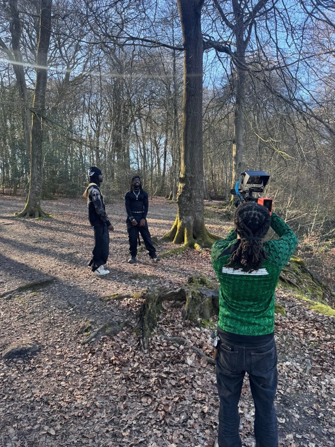 Two men standing outdoors in a forest, being filmed by a woman with a camera.
