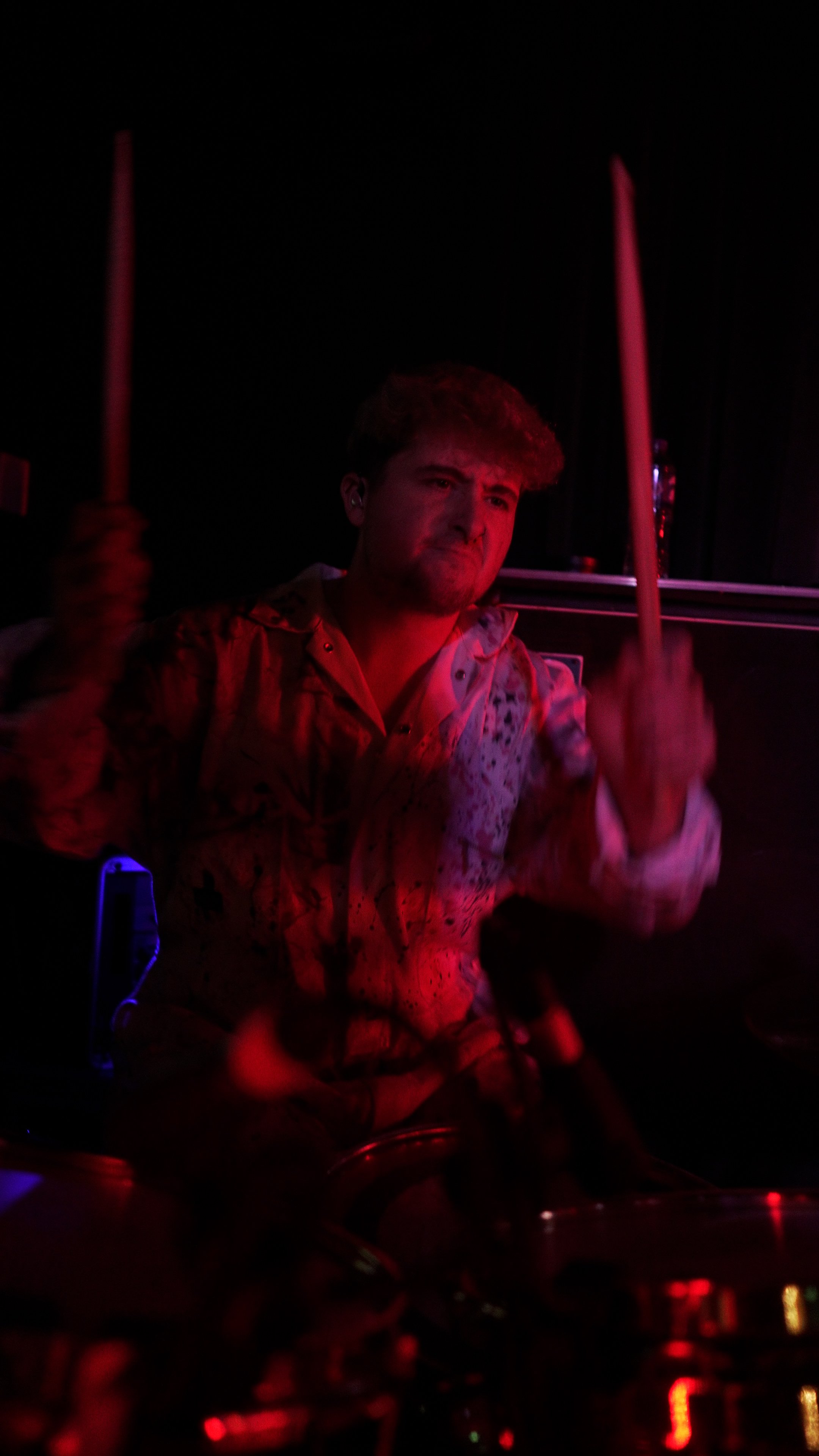 A young man playing drums on a dark stage illuminated by red and purple lights.