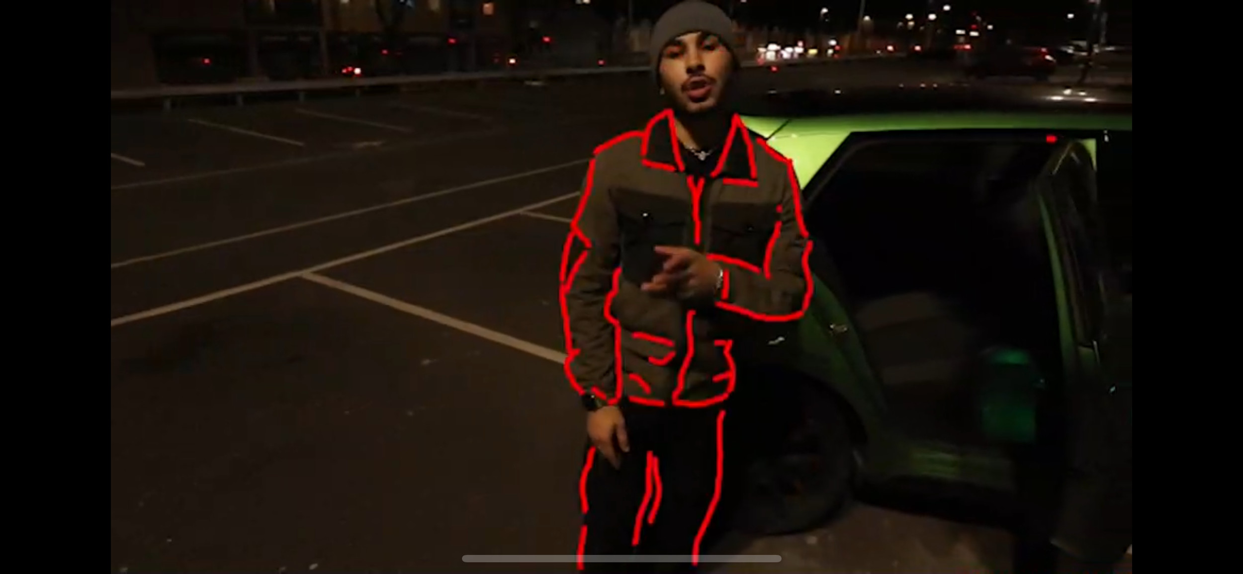 A young man standing next to a green vehicle in a parking lot at night, holding a phone, with red outline highlighting his jacket, pants, and arm.
