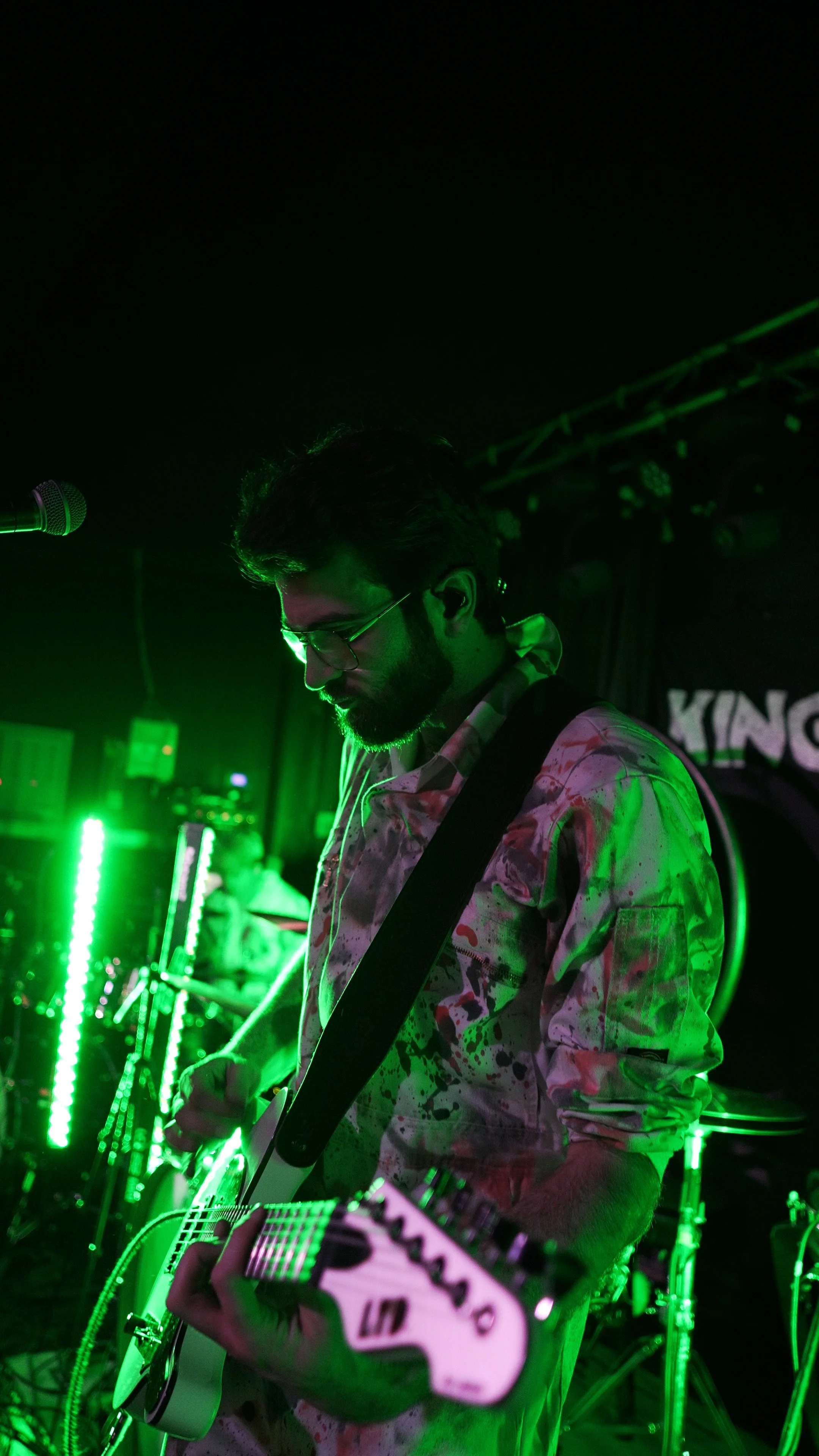A man with glasses playing an electric guitar on stage, illuminated by green stage lights, with other band members and equipment visible in the background.
