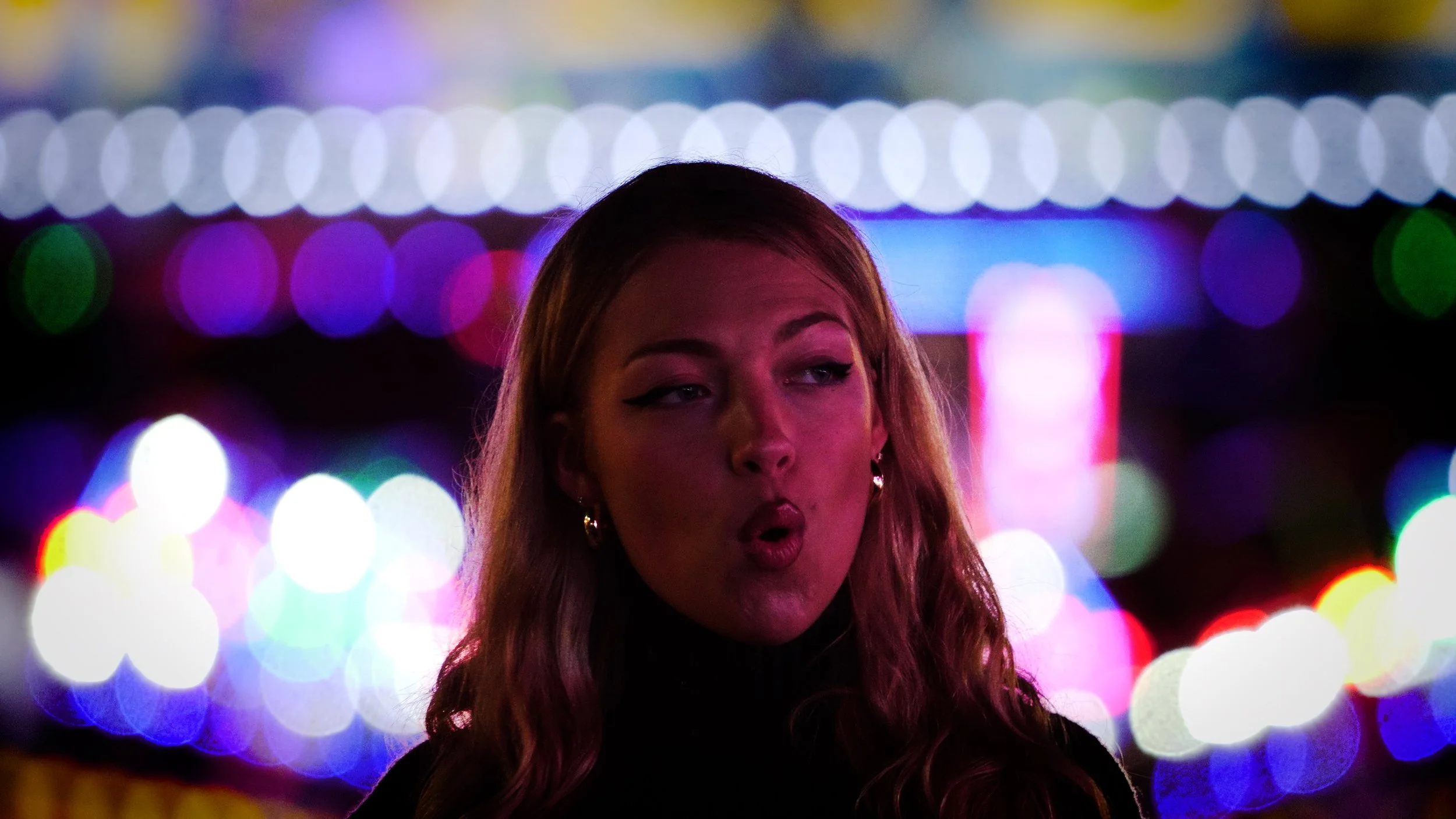 A young woman with long wavy hair and hoop earrings making a playful face in front of colorful bokeh lights.