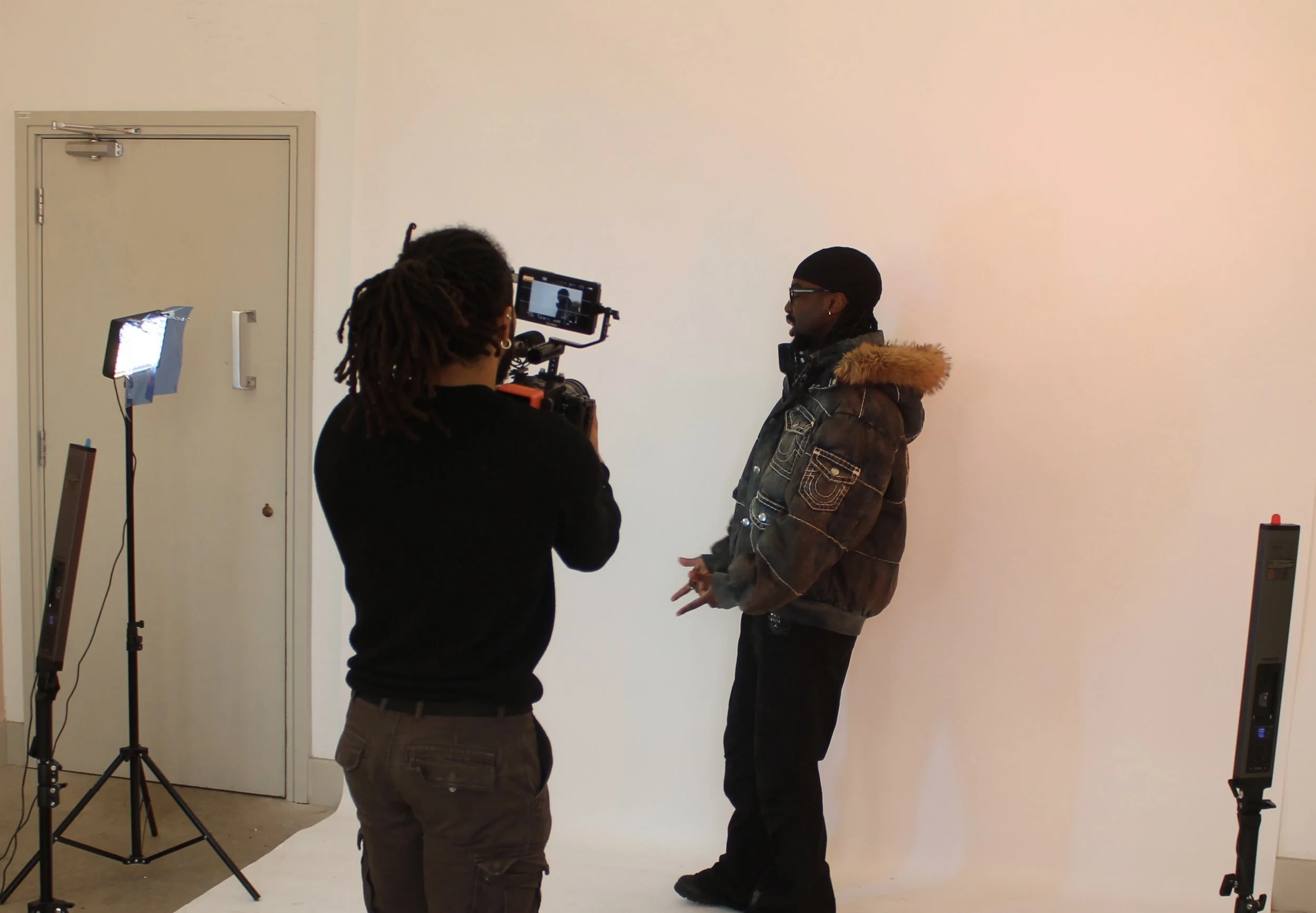 Behind the scenes of a photo shoot with two men, one holding a camera and the other posing, set up with lighting and a plain white backdrop.