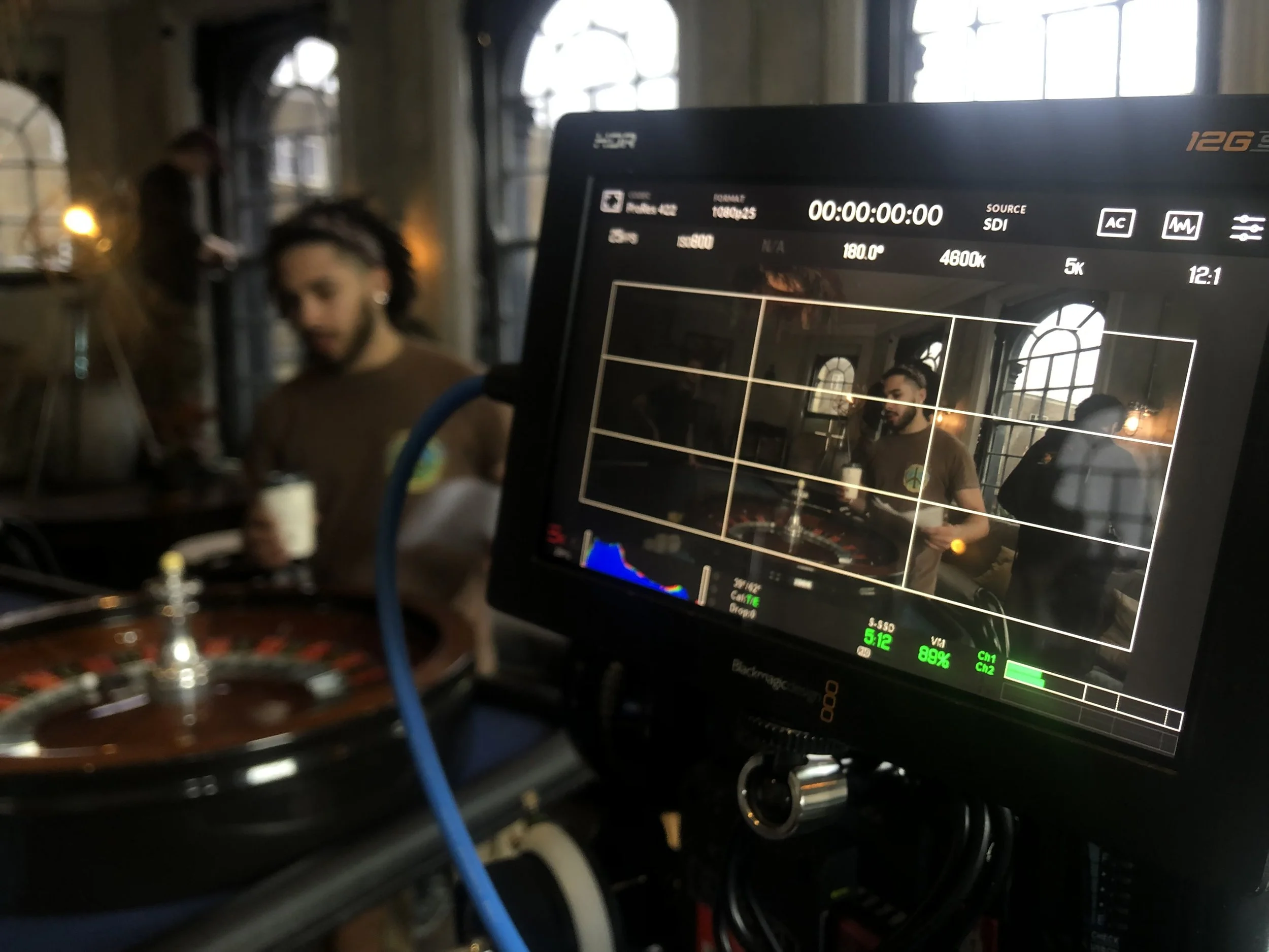 Film director's monitor showing a scene being shot in a dimly lit room with large arched windows and industrial-style decor, featuring a man with dark hair and a beard wearing a brown shirt, holding a coffee mug.