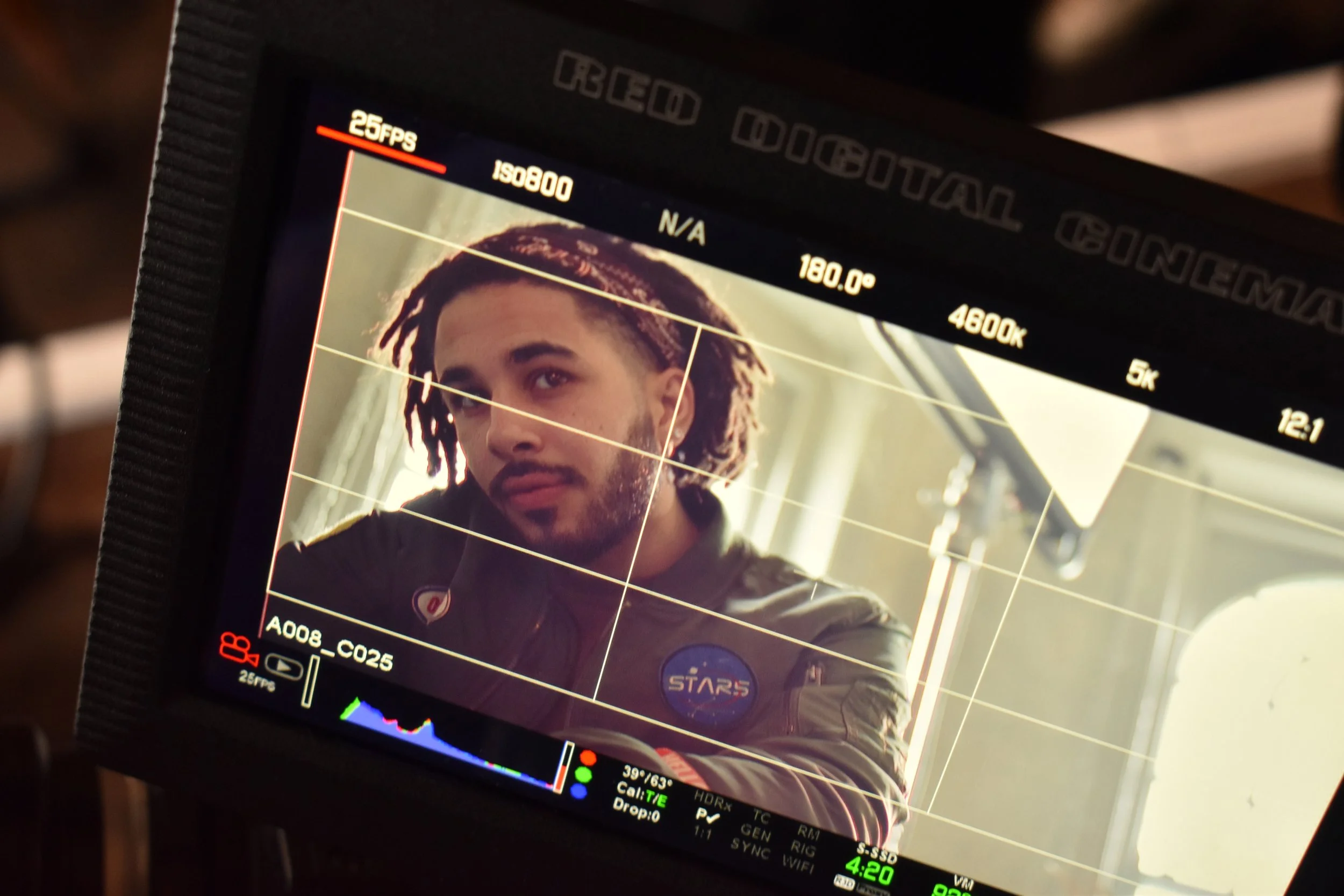 A camera monitor showing a man with dreadlocks and a beard in a space suit with a NASA badge, looking slightly to the side, in a well-lit room.