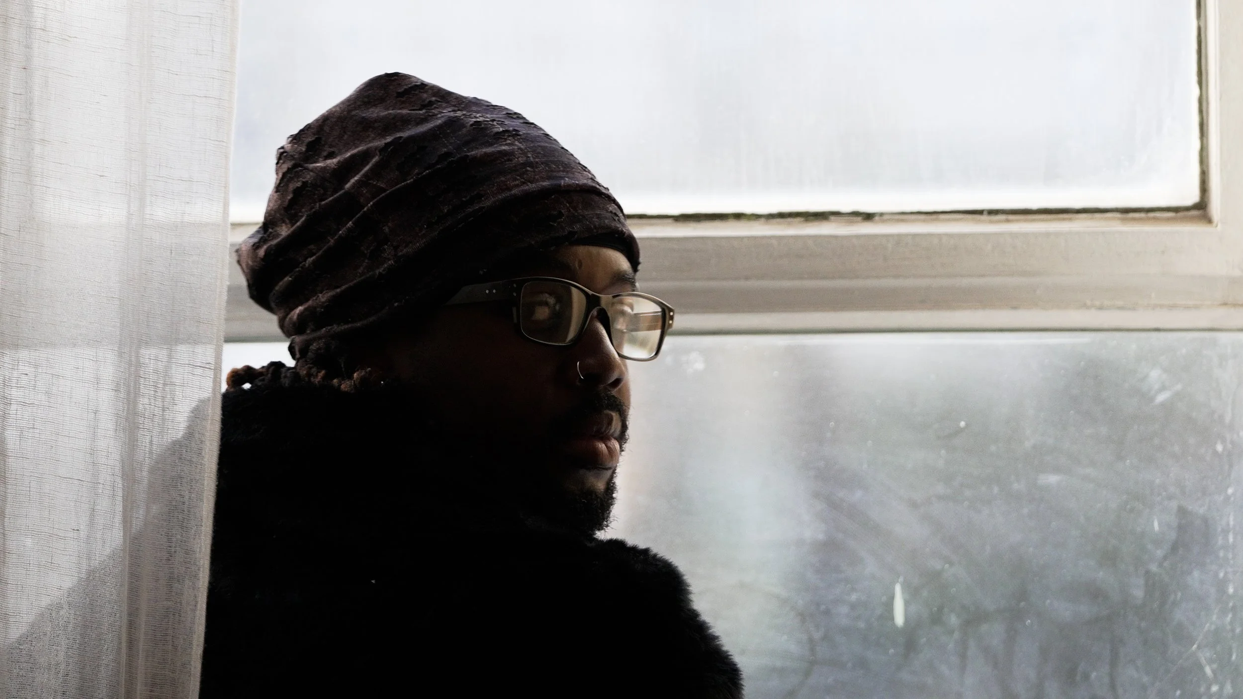 A person with glasses, a nose ring, and a headwrap, looking out a window.