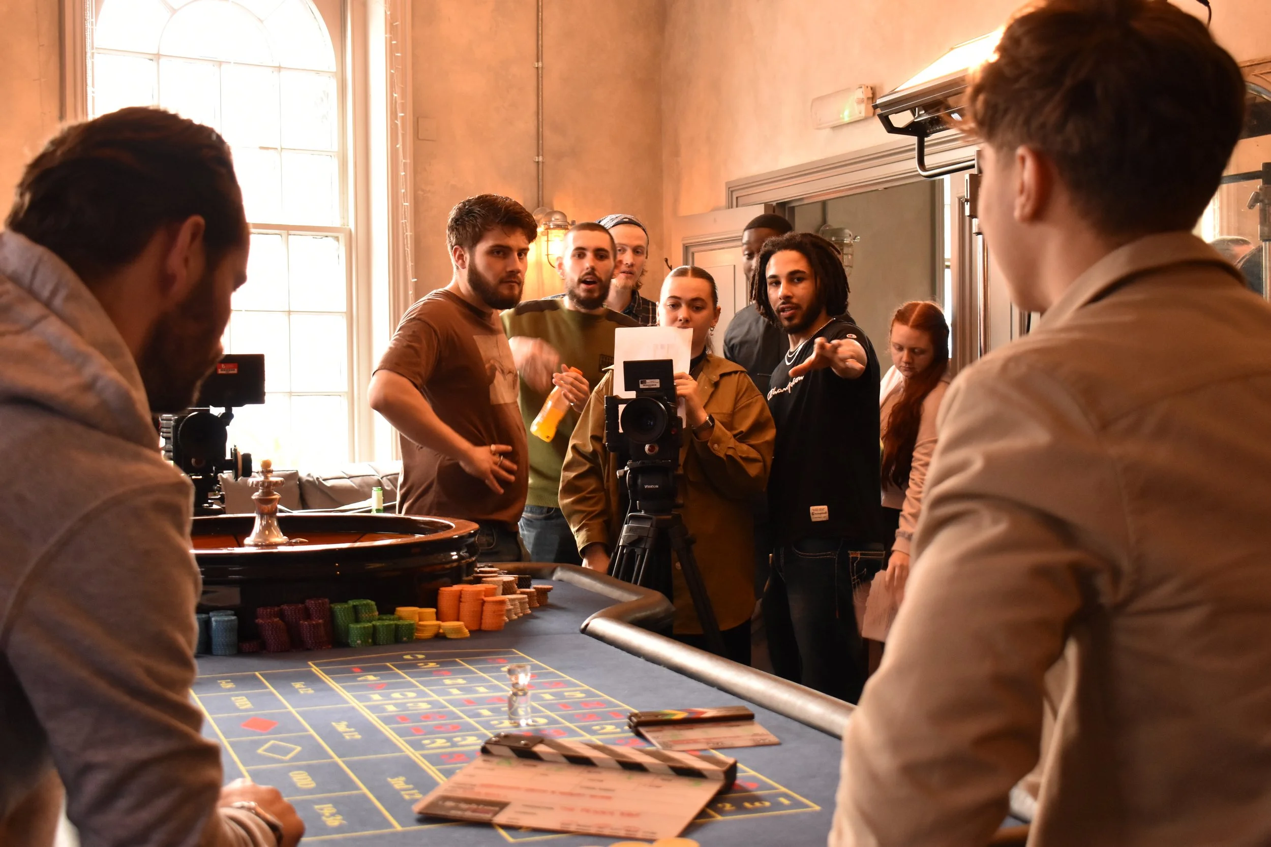 A group of people gathered around a roulette table in a casino or gambling setting. Several individuals are standing behind the table, some looking towards the camera and others engaging with or observing the game. The foreground shows two men seated at the roulette table, with chips and betting slips on the table. The room has large windows and warm lighting.