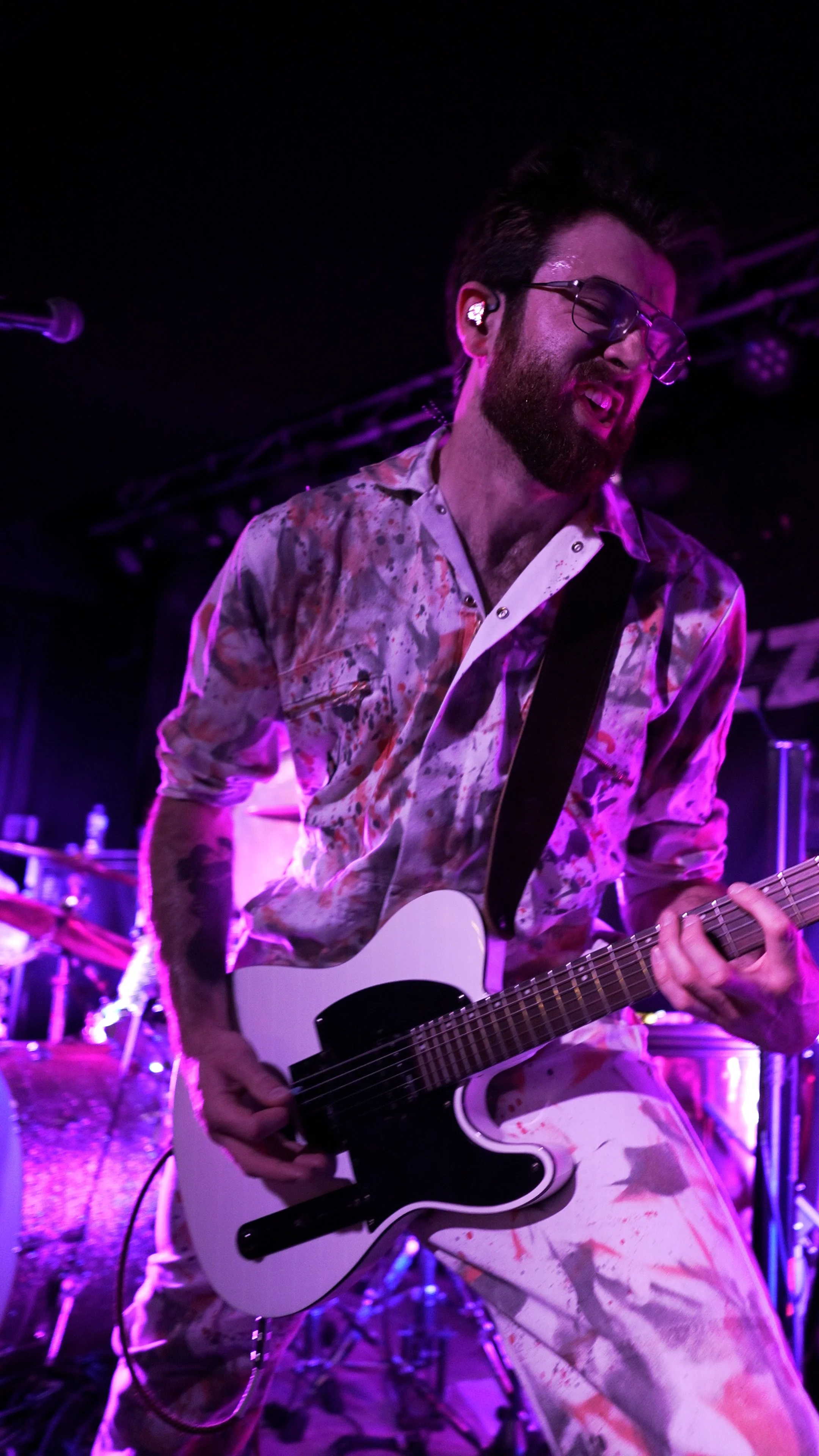 A male guitarist performing passionately on stage, wearing glasses and a paint-splattered shirt, with purple stage lighting.