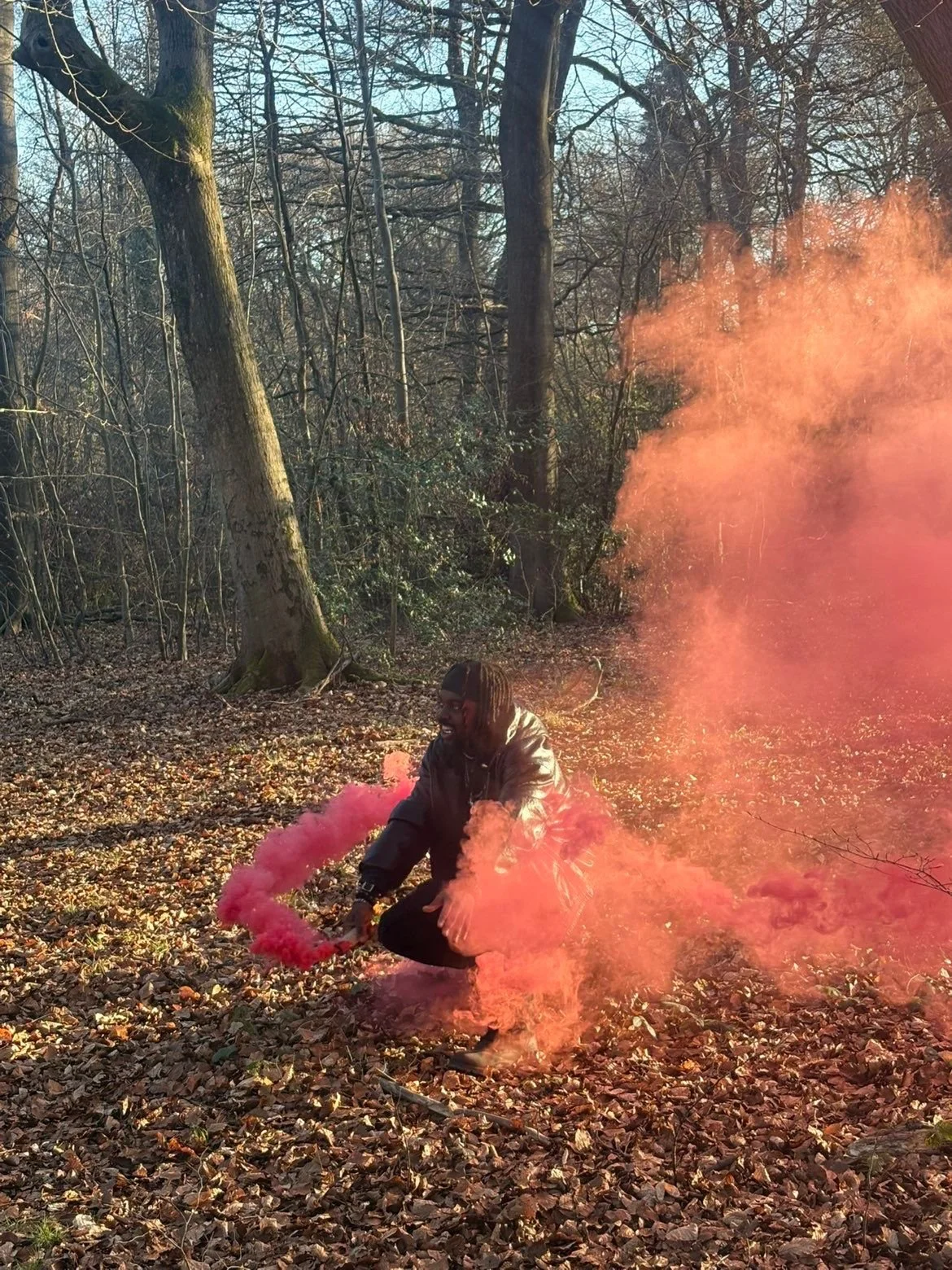 A person crouching on a forest floor covered in fallen leaves, holding a pink smoke flare, with pink smoke billowing around them amidst trees with bare branches.