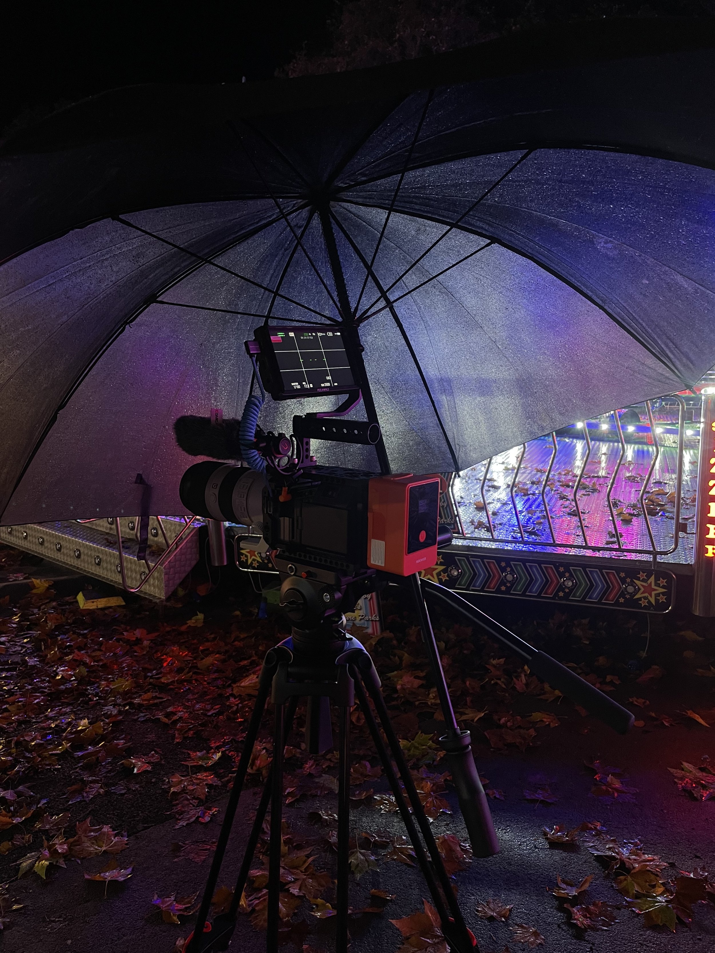 Video camera on tripod capturing footage under a wet, open umbrella at night with colorful carnival rides in the background and fallen autumn leaves on the ground.