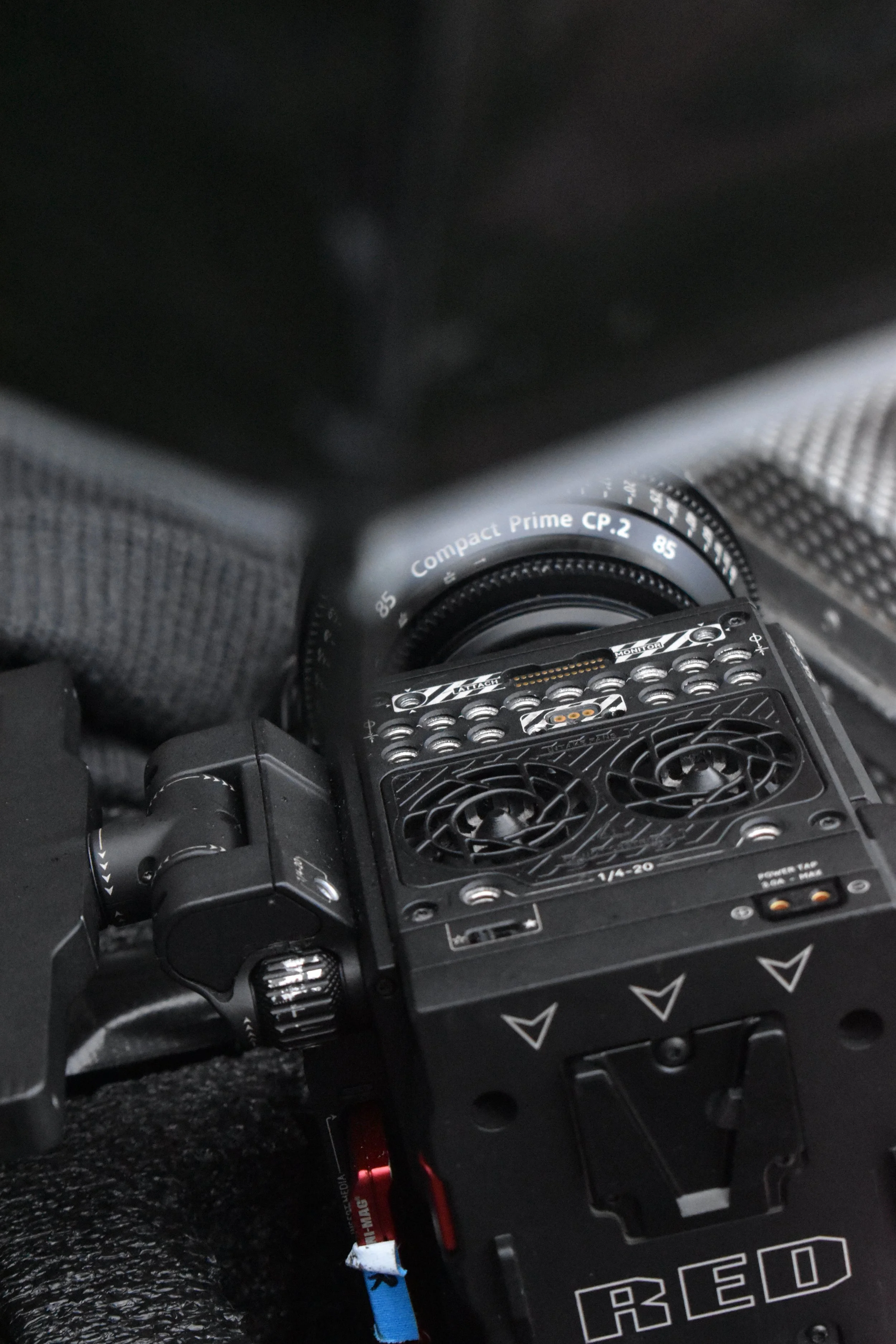 Close-up of a professional cinema camera with a black matte finish, attached to a Greg Black 85mm Compact Prime lens, placed on a textured surface.