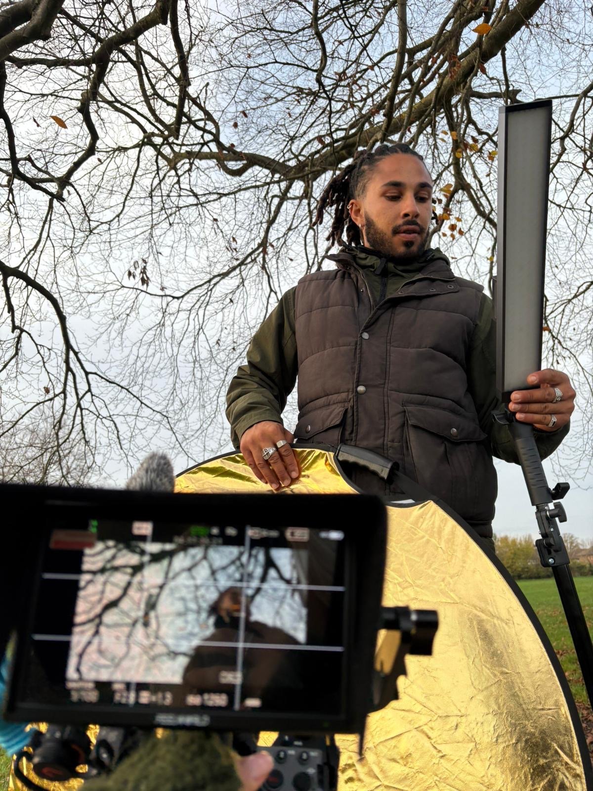 A man is holding a reflective gold disc outdoors with leafless trees and a cloudy sky in the background, while a camera on a tripod captures him. The scene appears to be part of a photoshoot or filming setup.