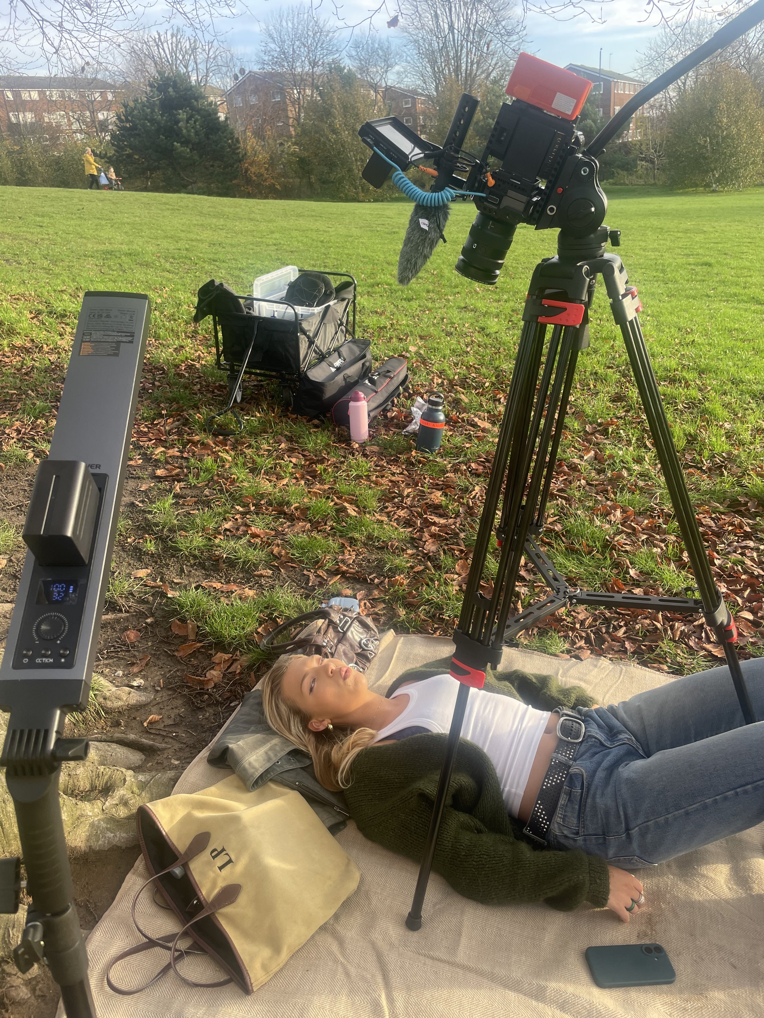 A woman lying on a blanket on the ground in a park, with camera and filming equipment set up around her, capturing a scene outdoors.