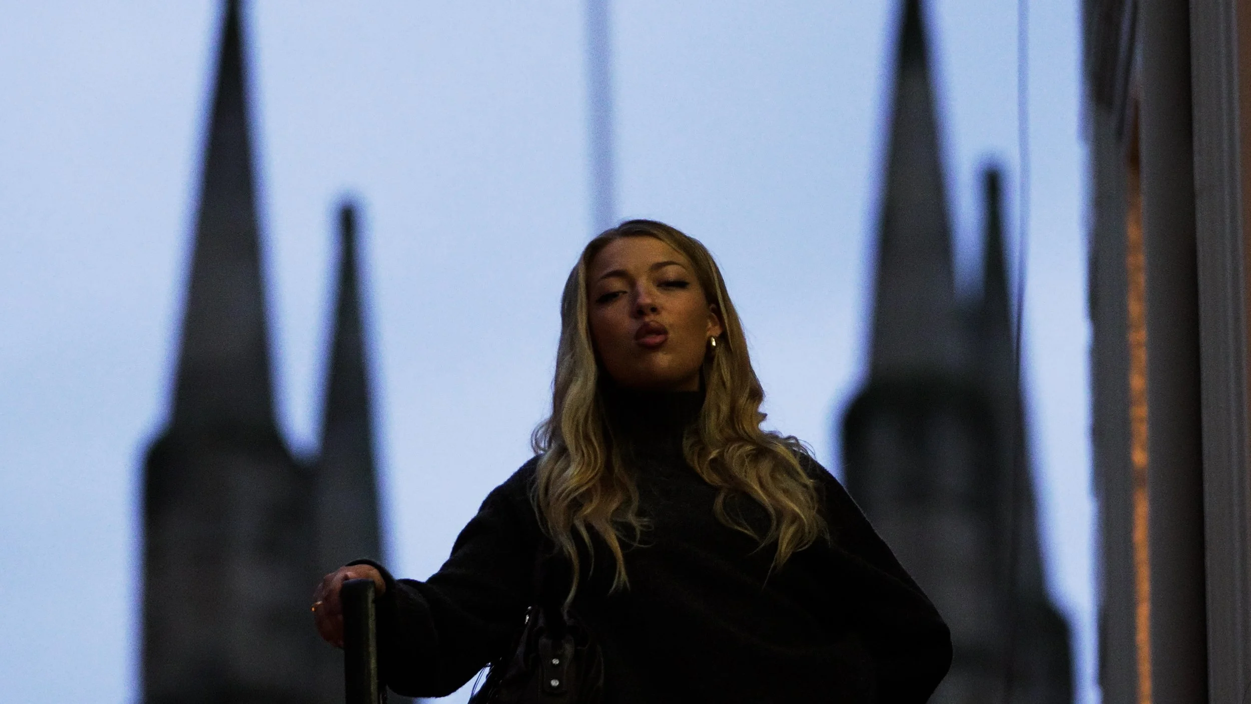 A woman with long blonde hair posing with her lips puckered, standing between tall dark spires against a cloudy sky.