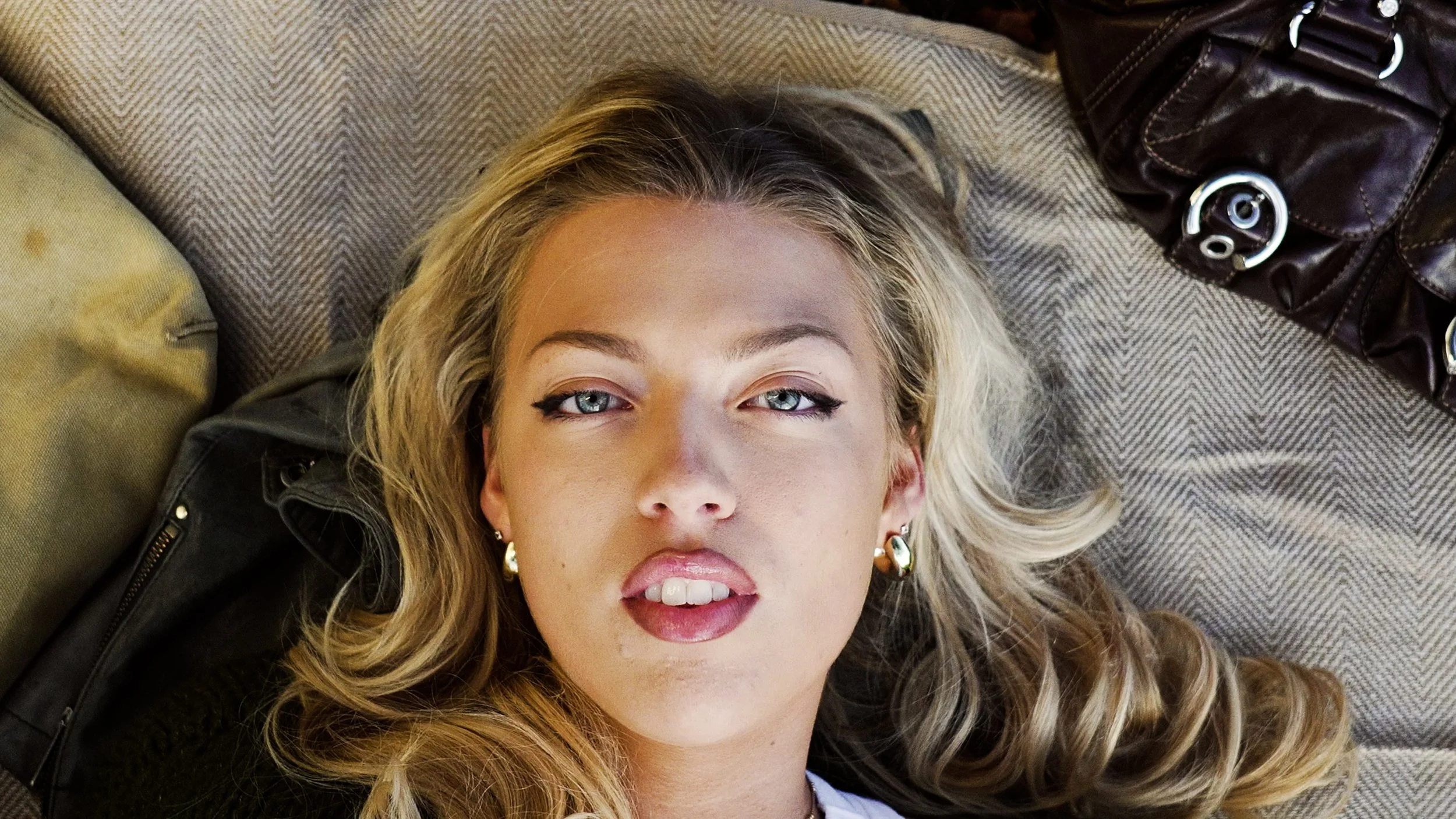 A woman with blonde wavy hair, blue eyes, and makeup, lying on a couch or bed, looking at the camera, with a leather handbag and a beige pillow nearby.