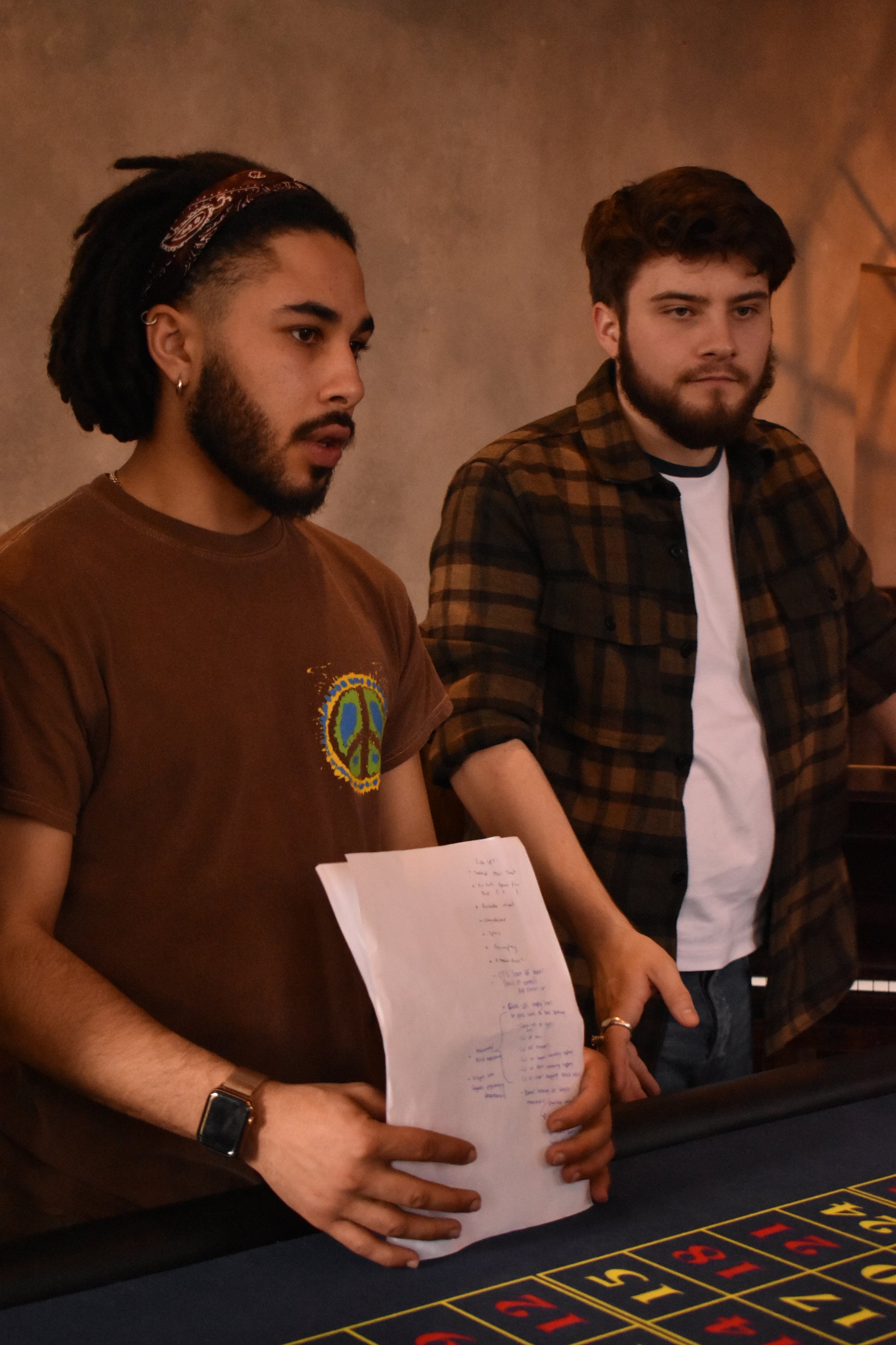 Two men standing at a roulette table in a casino or gaming room, looking focused. The man on the left has dreadlocks, a bandana, a brown t-shirt with a peace sign, and a smartwatch. He holds a notebook or sheet of paper. The man on the right has brown hair, a beard, and is wearing a brown checkered shirt over a white t-shirt.