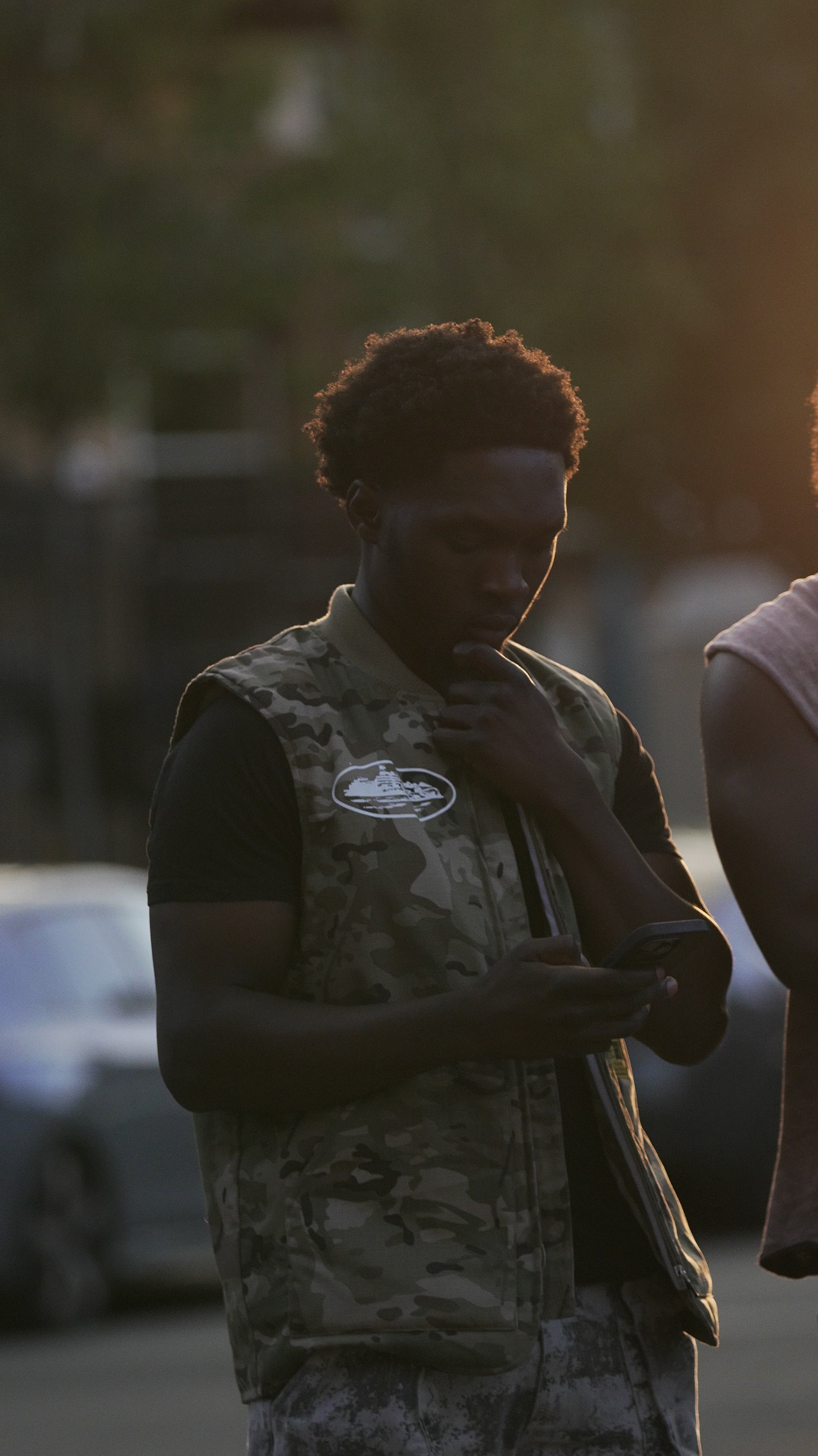 Young man with curly hair wearing a camouflage vest and black t-shirt, looking at his phone outdoors during sunset.