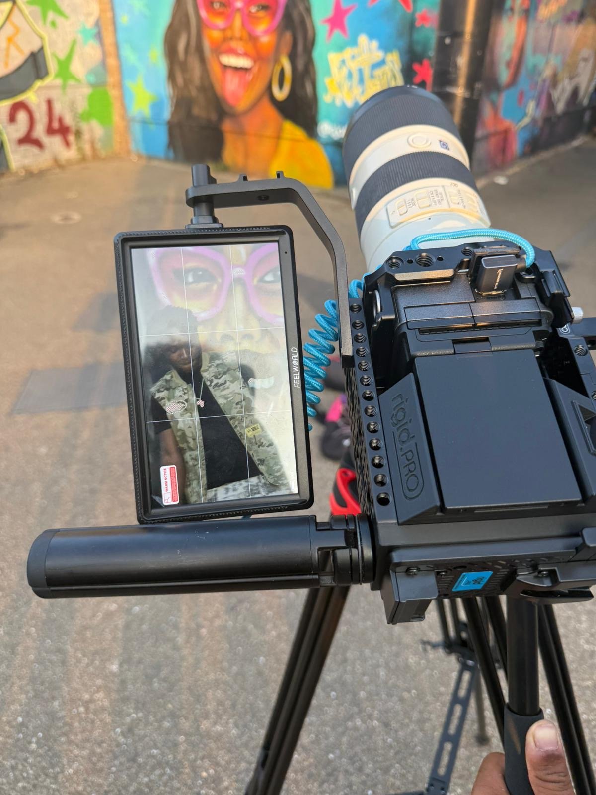 The photograph shows a professional camera mounted on a tripod, focusing on a colorful mural of a woman with glasses and earrings. The mural features vibrant colors and various artistic details, with a person taking a picture of the mural with a camera.