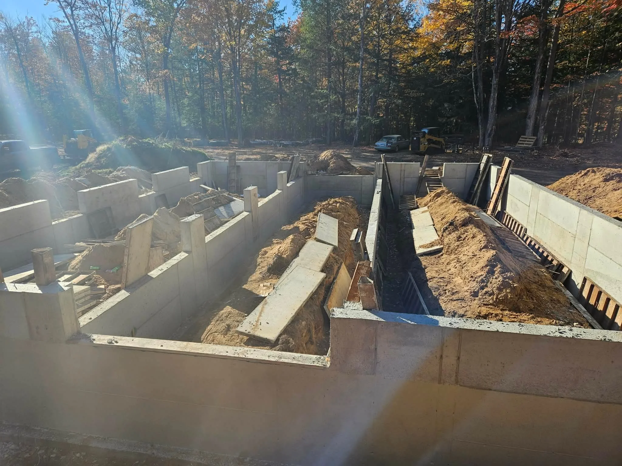 Construction site with concrete foundation walls and excavation areas surrounded by dirt, with trees and vehicles in the background.