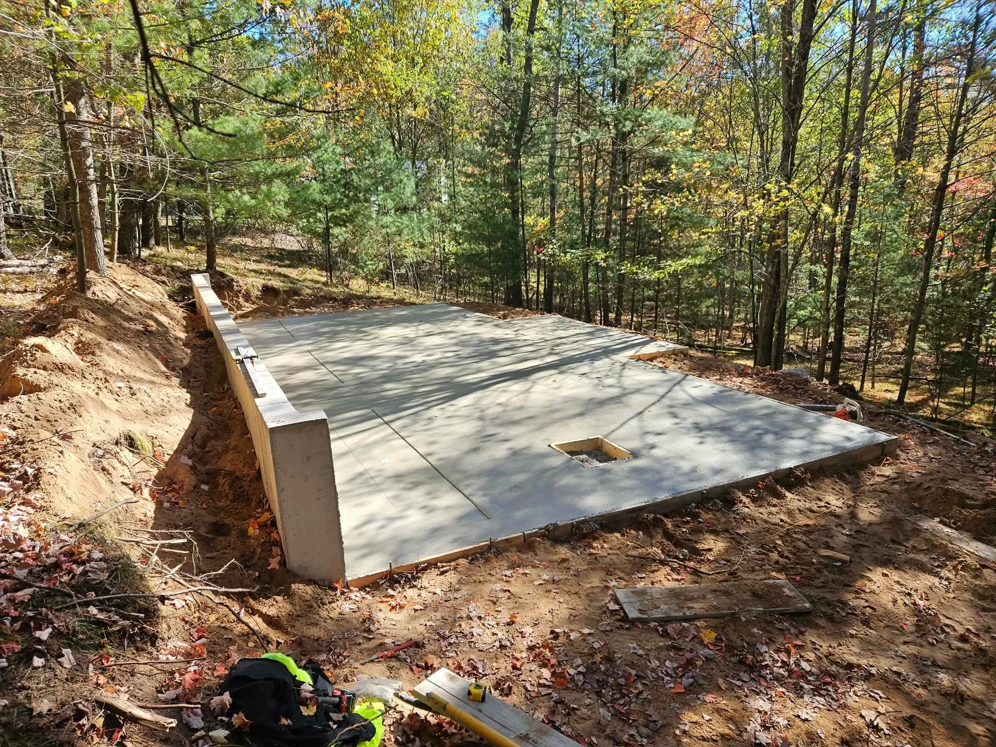 Concrete foundation slab on a dirt clearing surrounded by trees in a wooded area.