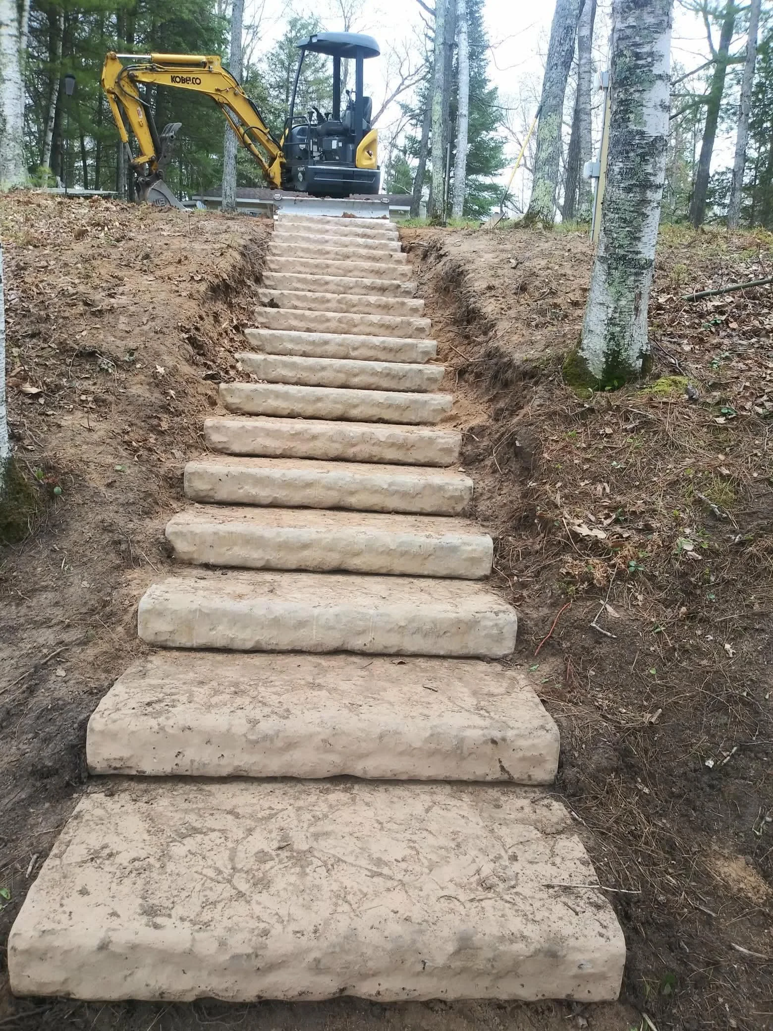 Concrete steps leading up a hill with a small yellow excavator at the top in a wooded area.