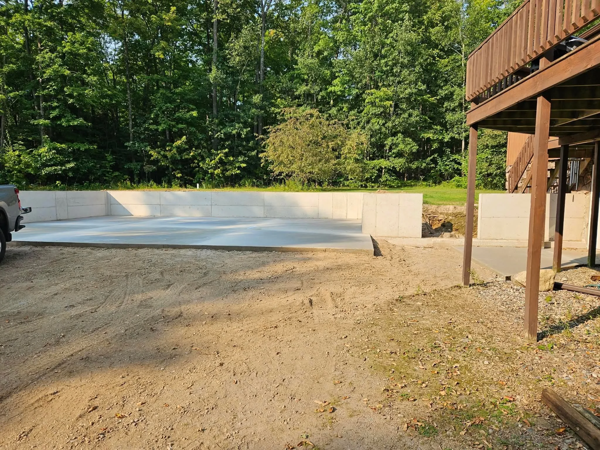 An outdoor construction site with a partially built concrete pool, a pickup truck on the left, a wooded background, and a wooden deck structure on the right.