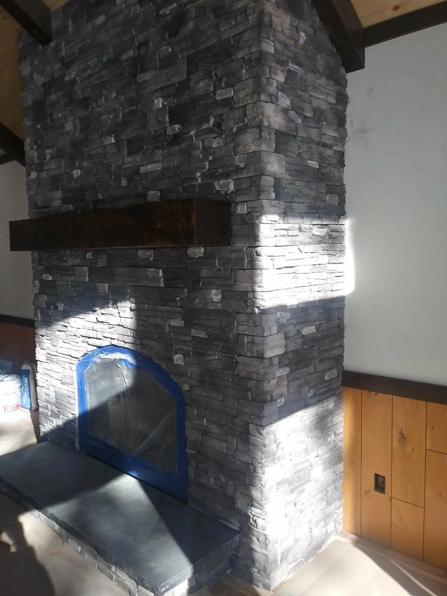 A stone fireplace with a wooden mantel and a covered firebox opening, built with dark gray stacked stones, in a room with wood paneling on the lower wall.
