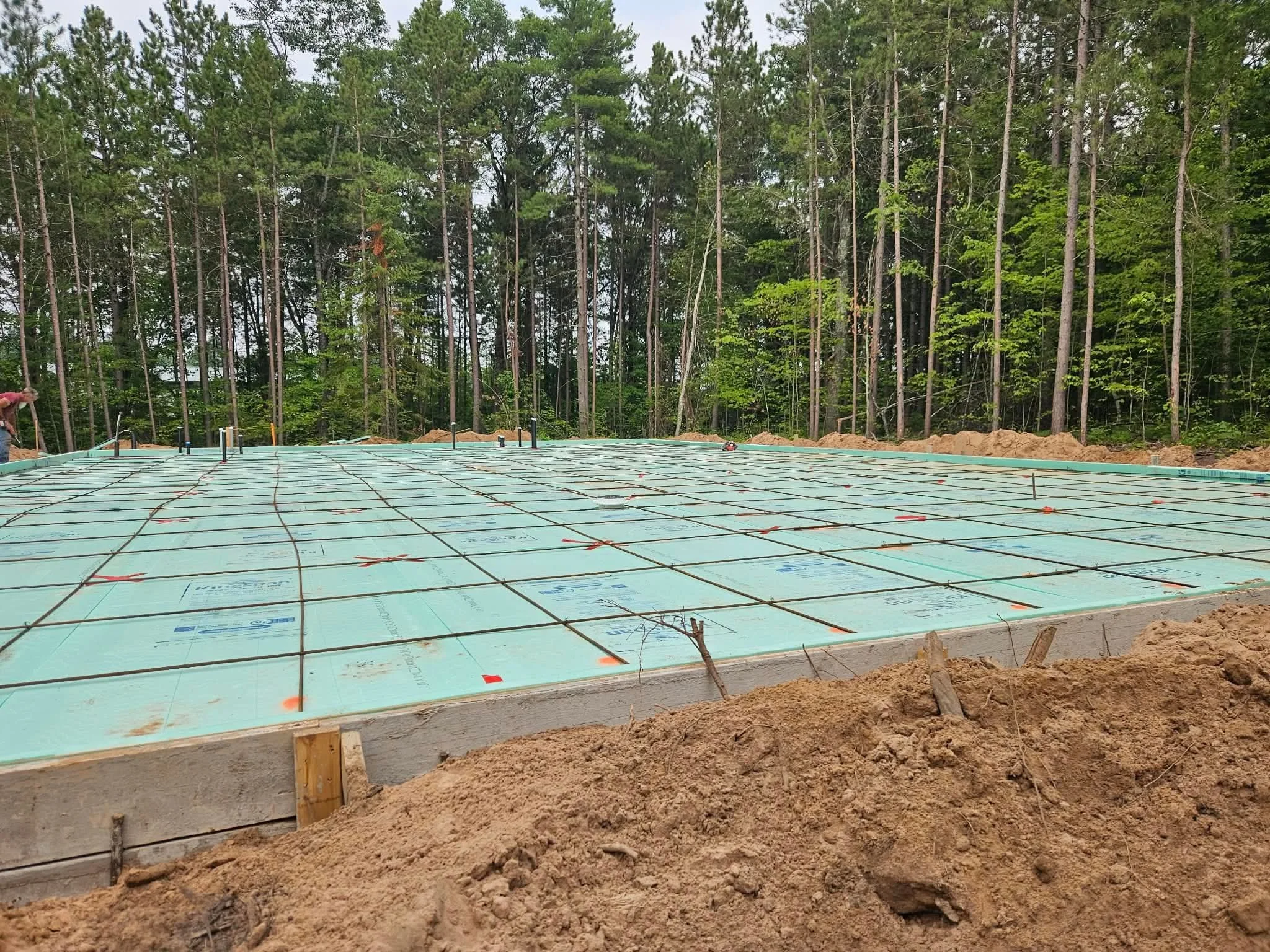 Construction site with a concrete foundation, green insulation, and rebar grid, surrounded by soil and trees in the background.