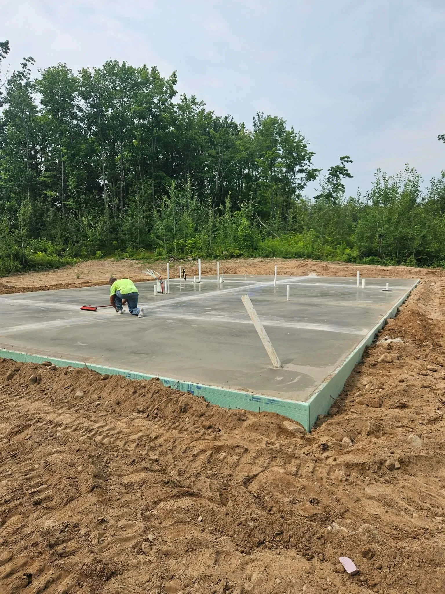 Construction worker working on a new foundation with plumbing pipes on a concrete slab in a cleared area surrounded by trees.
