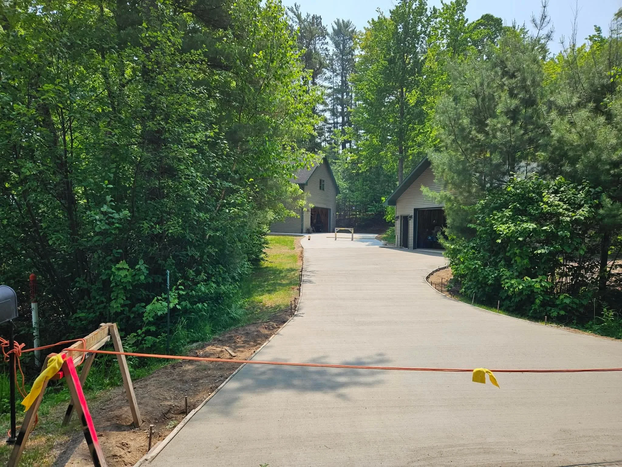 Recently poured concrete driveway leading to a house garage, surrounded by green trees and shrubs, with a construction barrier and equipment at the edge.
