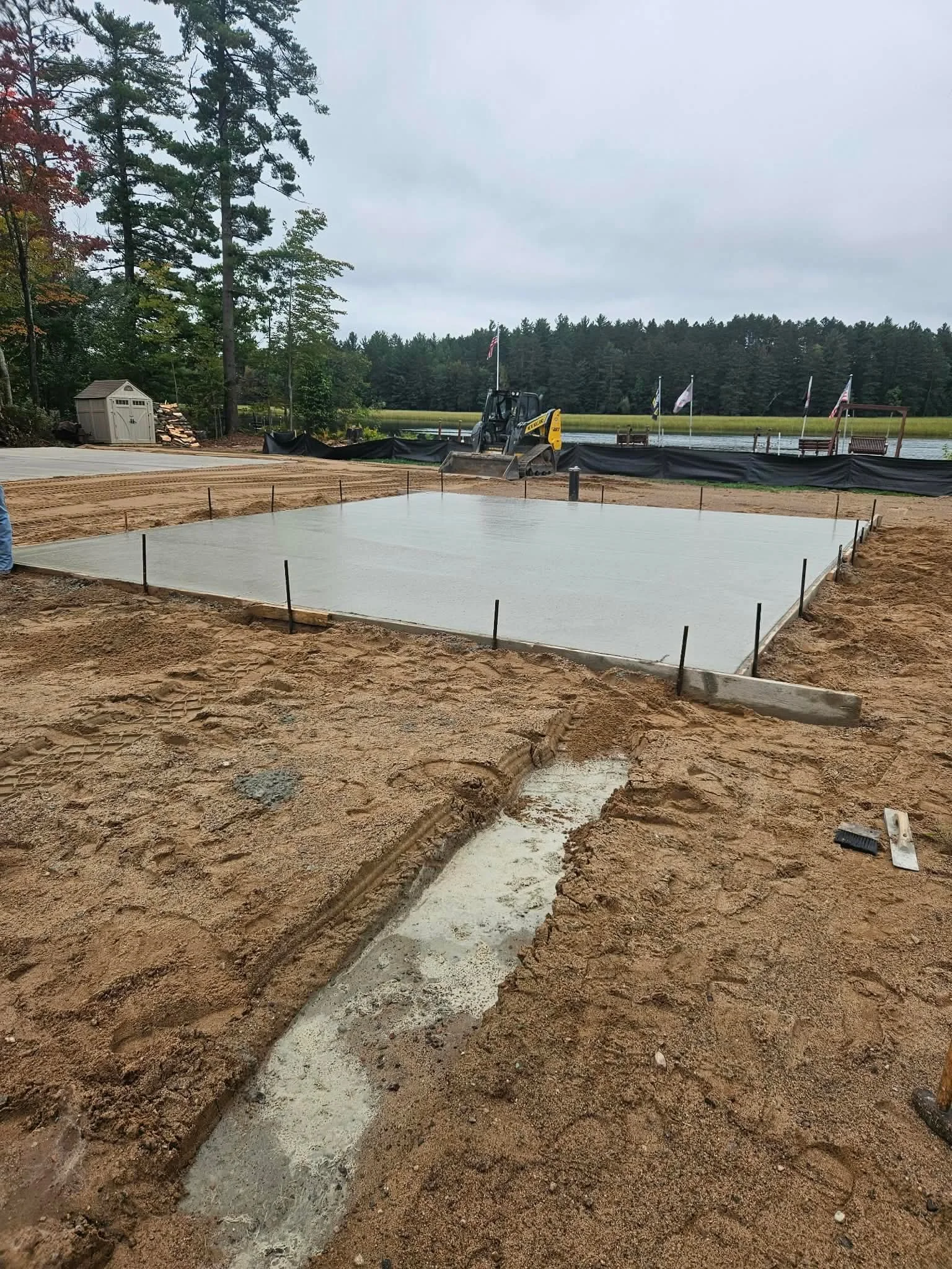 Construction site with freshly poured concrete slab, surrounded by dirt and marked with stakes, near a lake with flags and trees in the background.