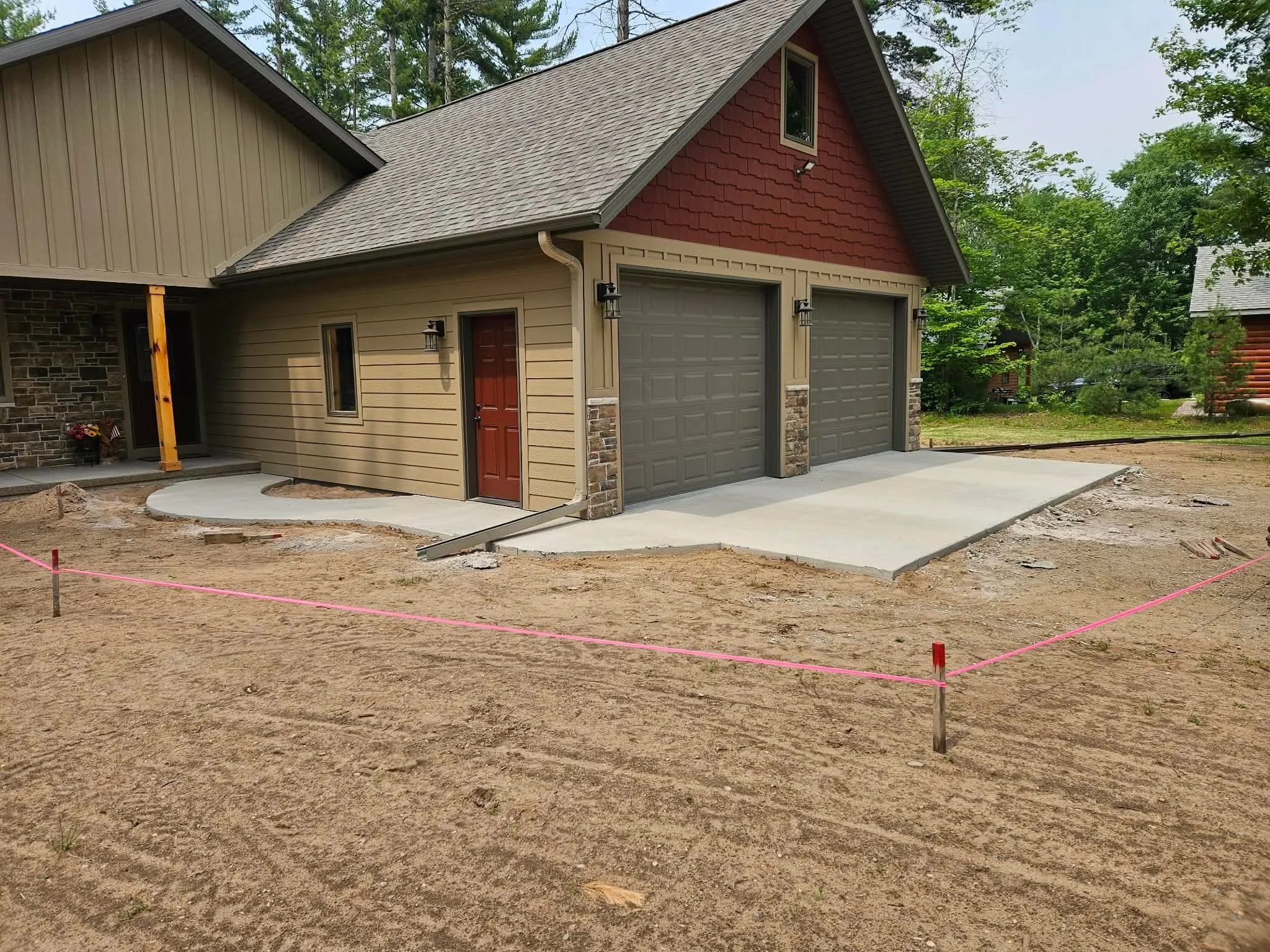 Newly constructed garage with double doors and a smaller side door, attached to a house with beige and red siding. The area in front of the garage is prepared for paving with a concrete driveway, and pink construction markers outline the section.