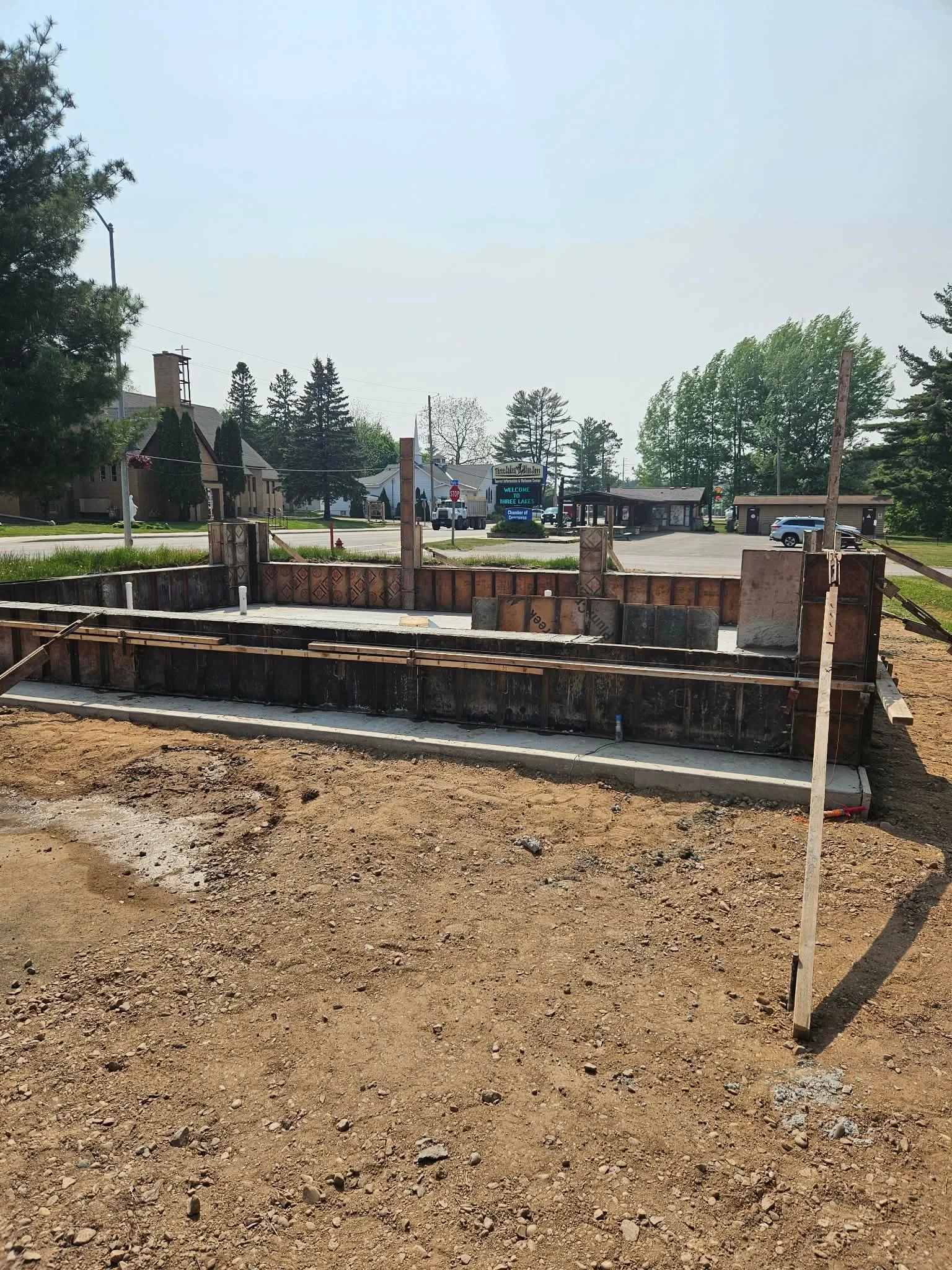Construction site with a concrete foundation and wooden formwork for a building, with residential houses and trees in the background.
