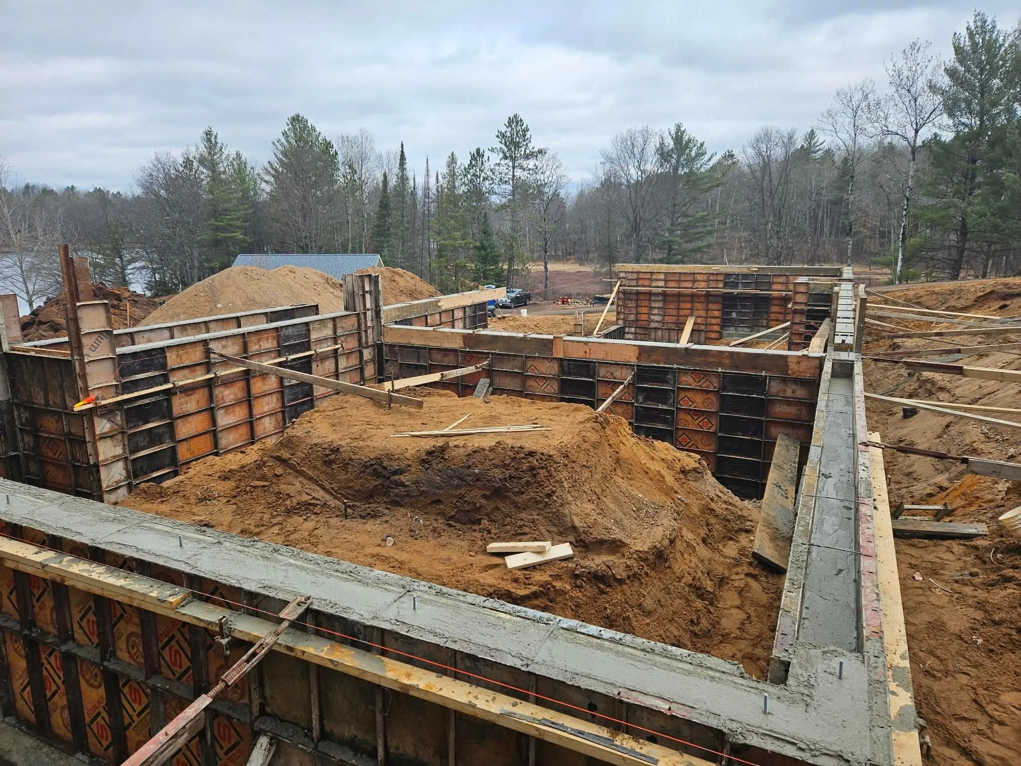 Construction site with concrete foundation and formwork for building walls, surrounded by dirt piles and outdoor trees under cloudy sky.