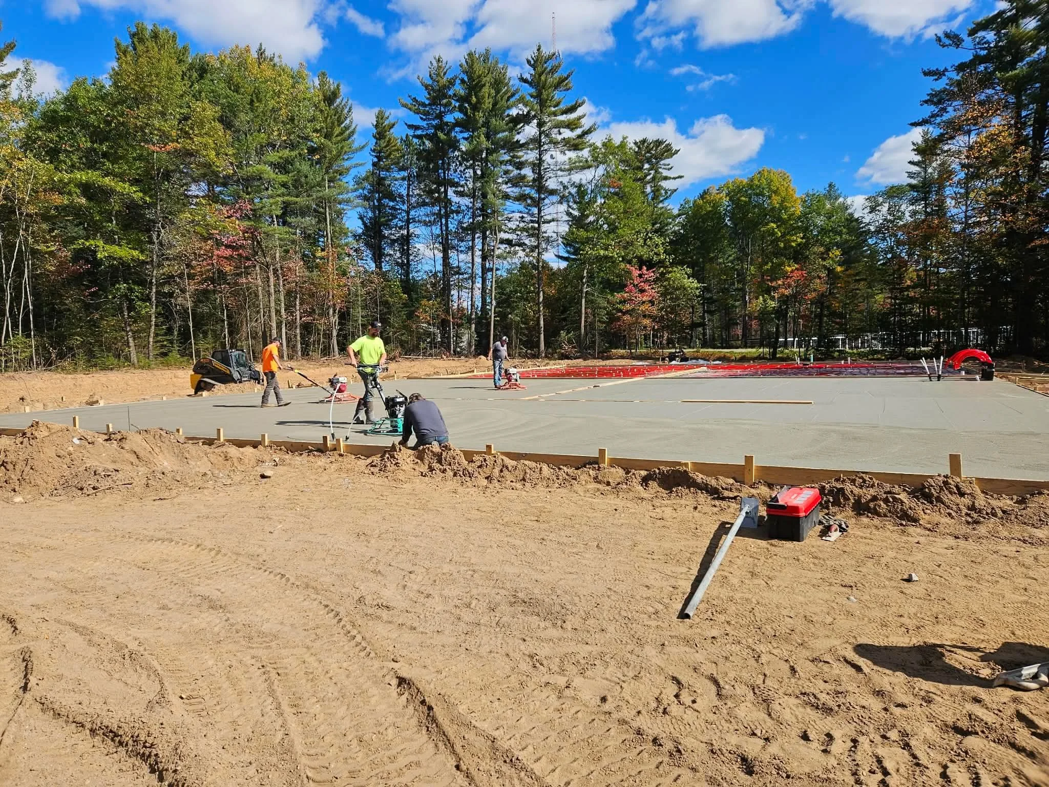 Construction workers flatten and prepare a concrete surface for a building foundation in a wooded area on a sunny day.