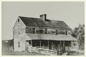 An old two-story wooden building with a steep roof and a chimney, supported on stilts with a porch and a wooden staircase leading to the entrance.