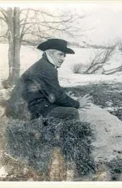 A person sitting outdoors near a body of water, wearing a dark coat and wide-brimmed hat, with leafless trees in the background.