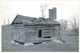 Old, dilapidated wooden cabin with missing roof and broken windows surrounded by leafless trees.