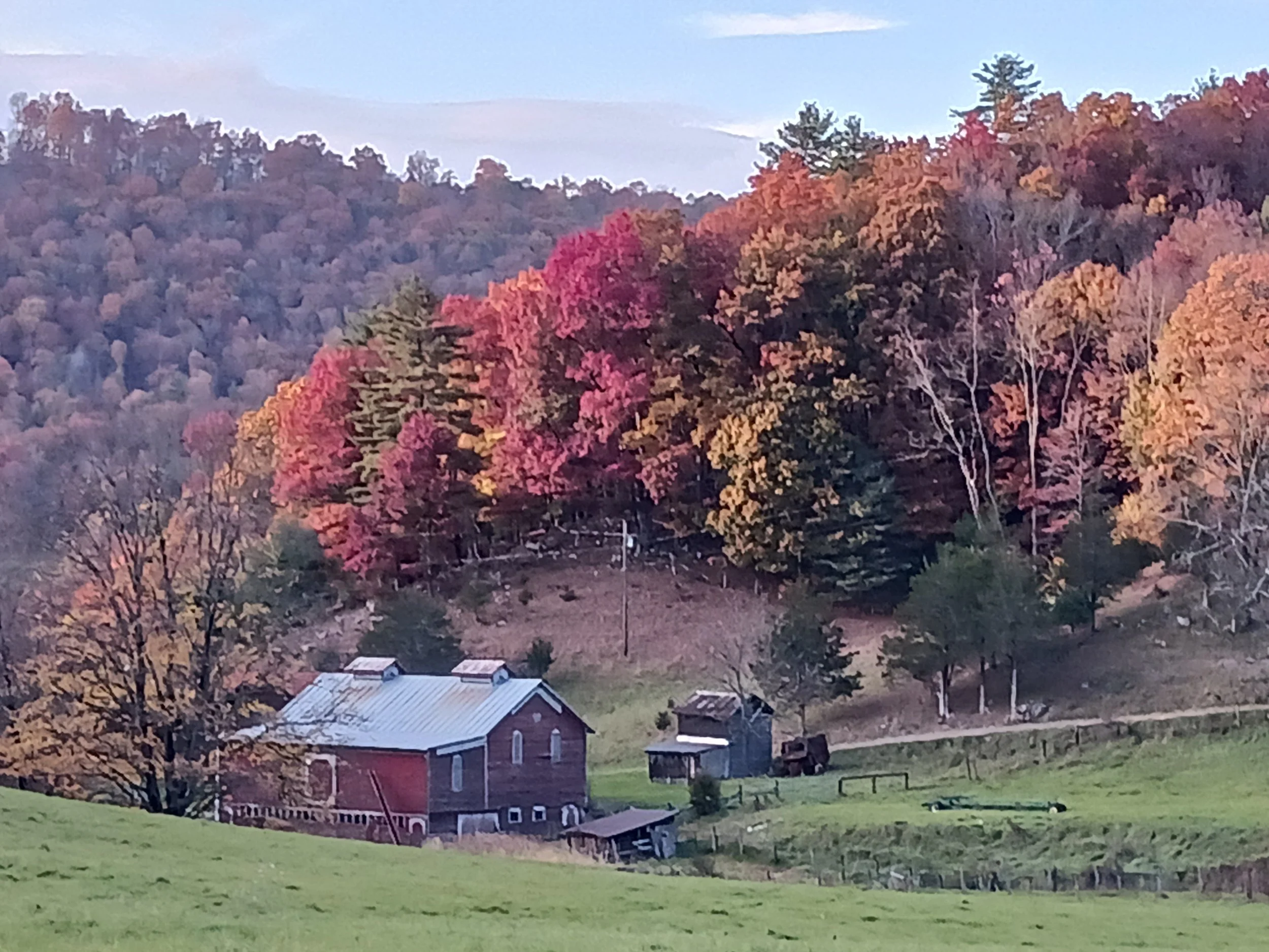 A rural scene with a red barn, a small shed, and a green field in the foreground, with autumn foliage covering a hillside in the background.